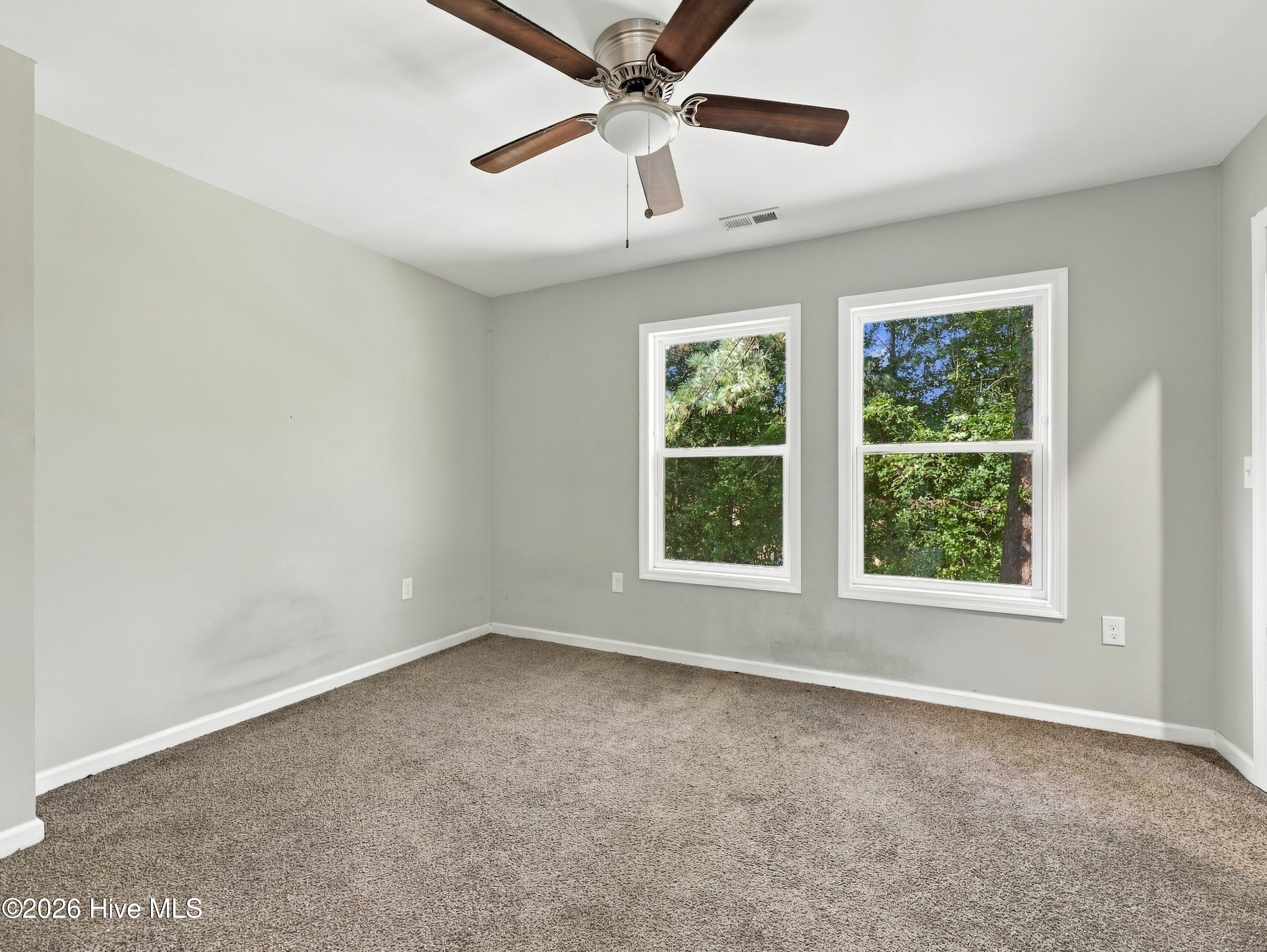 Empty bedroom with beige carpet, ceiling fan, and two windows overlooking trees.