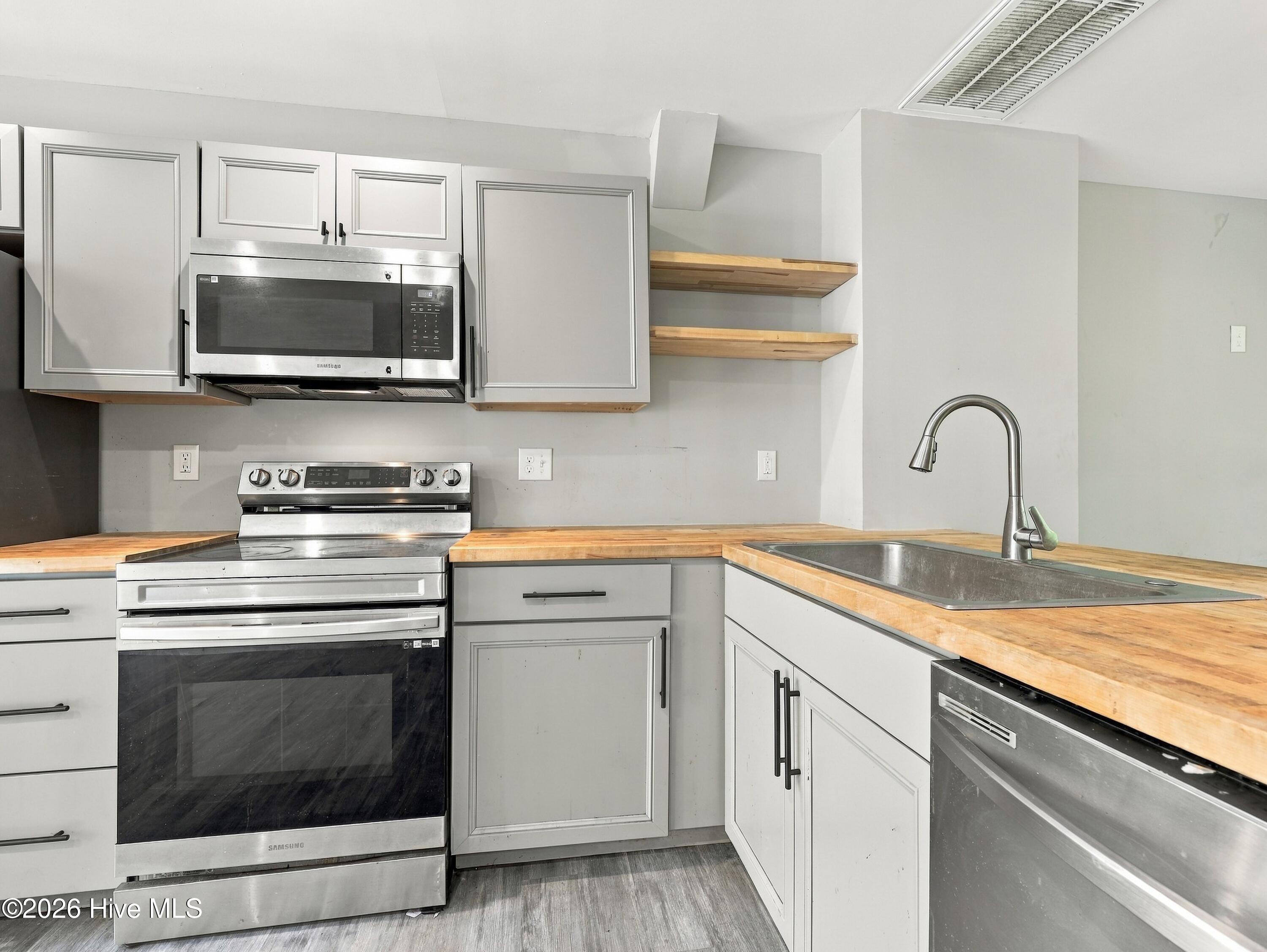 Kitchen with gray cabinets, butcher-block counters, stainless appliances, and open wood shelves.