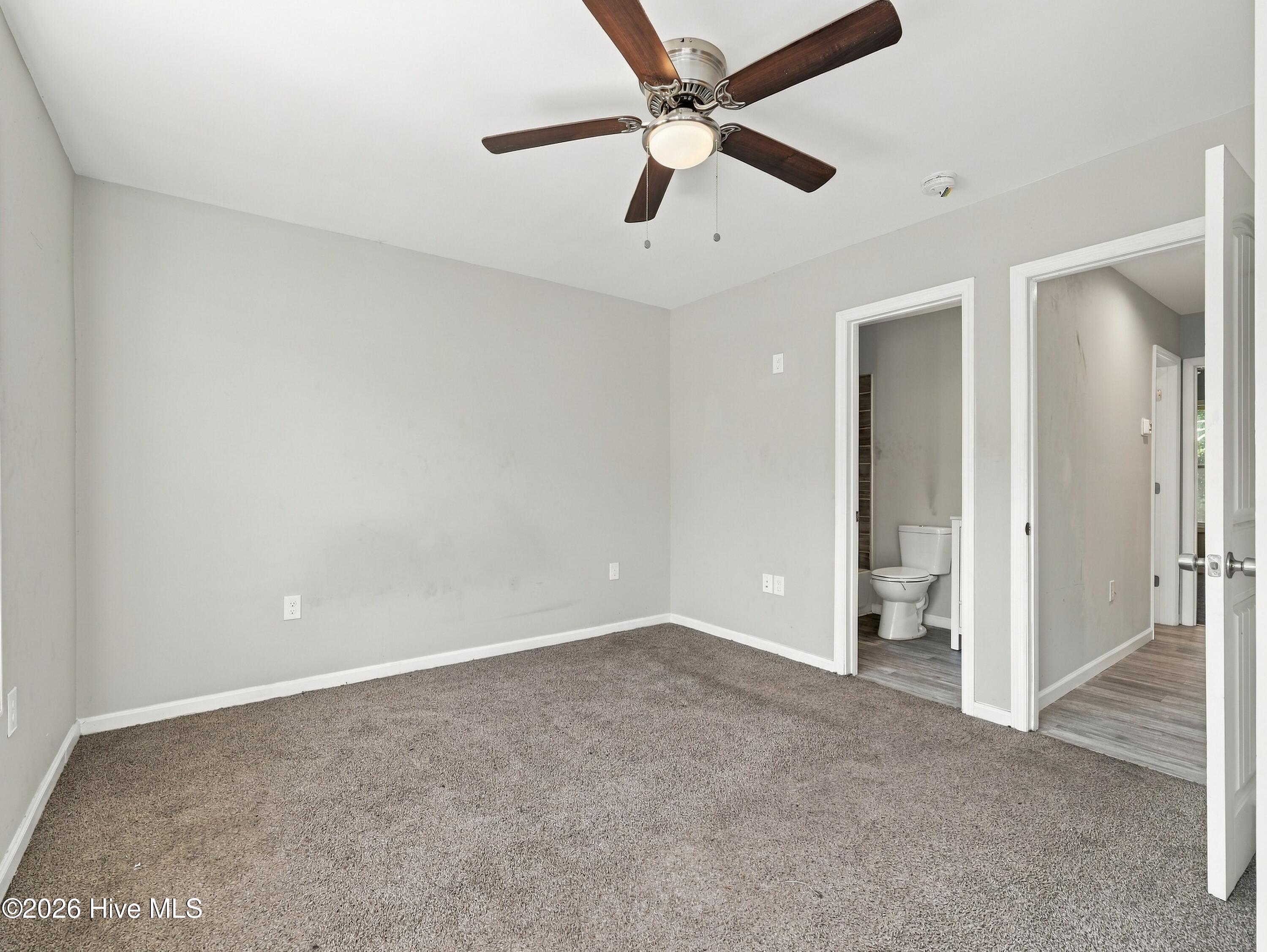 Empty bedroom with gray carpet, a ceiling fan, and open doorways to a bathroom and hall.