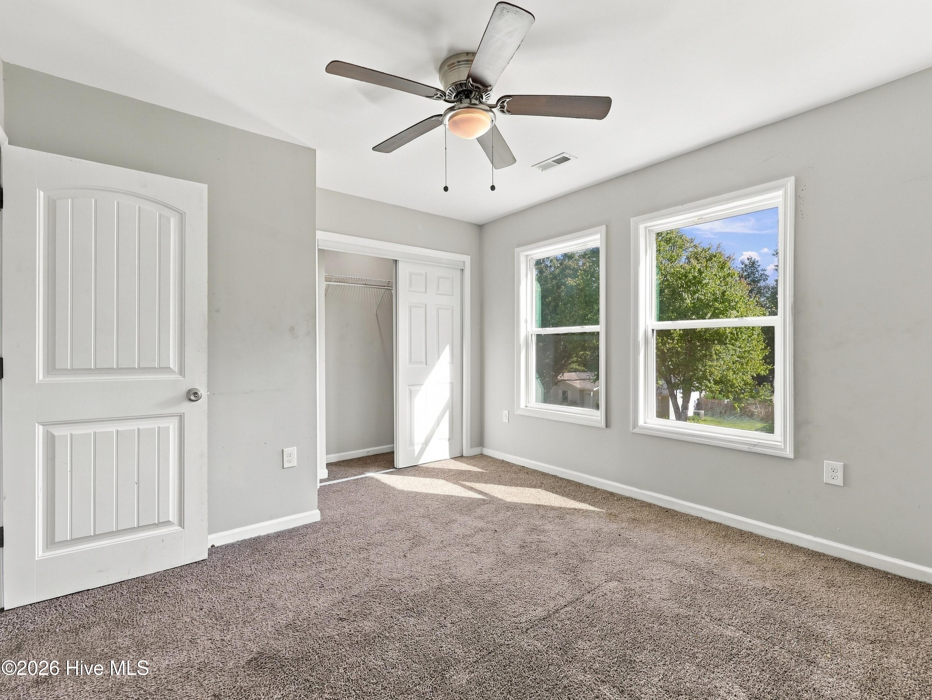 Empty bedroom with beige carpet, a ceiling fan, two windows, and an open closet.