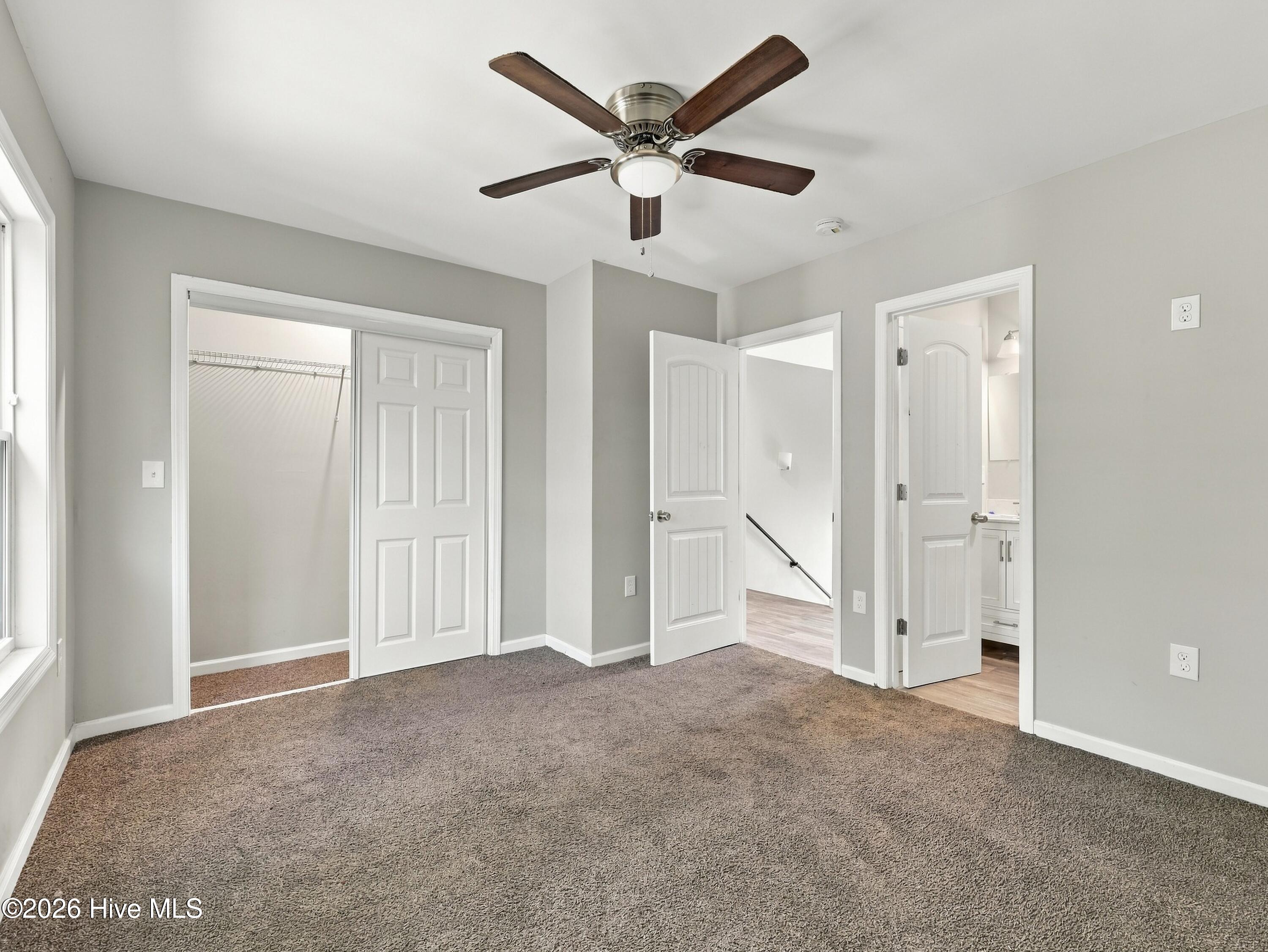 Empty bedroom with brown carpet, ceiling fan, and open closet and bathroom doorways.