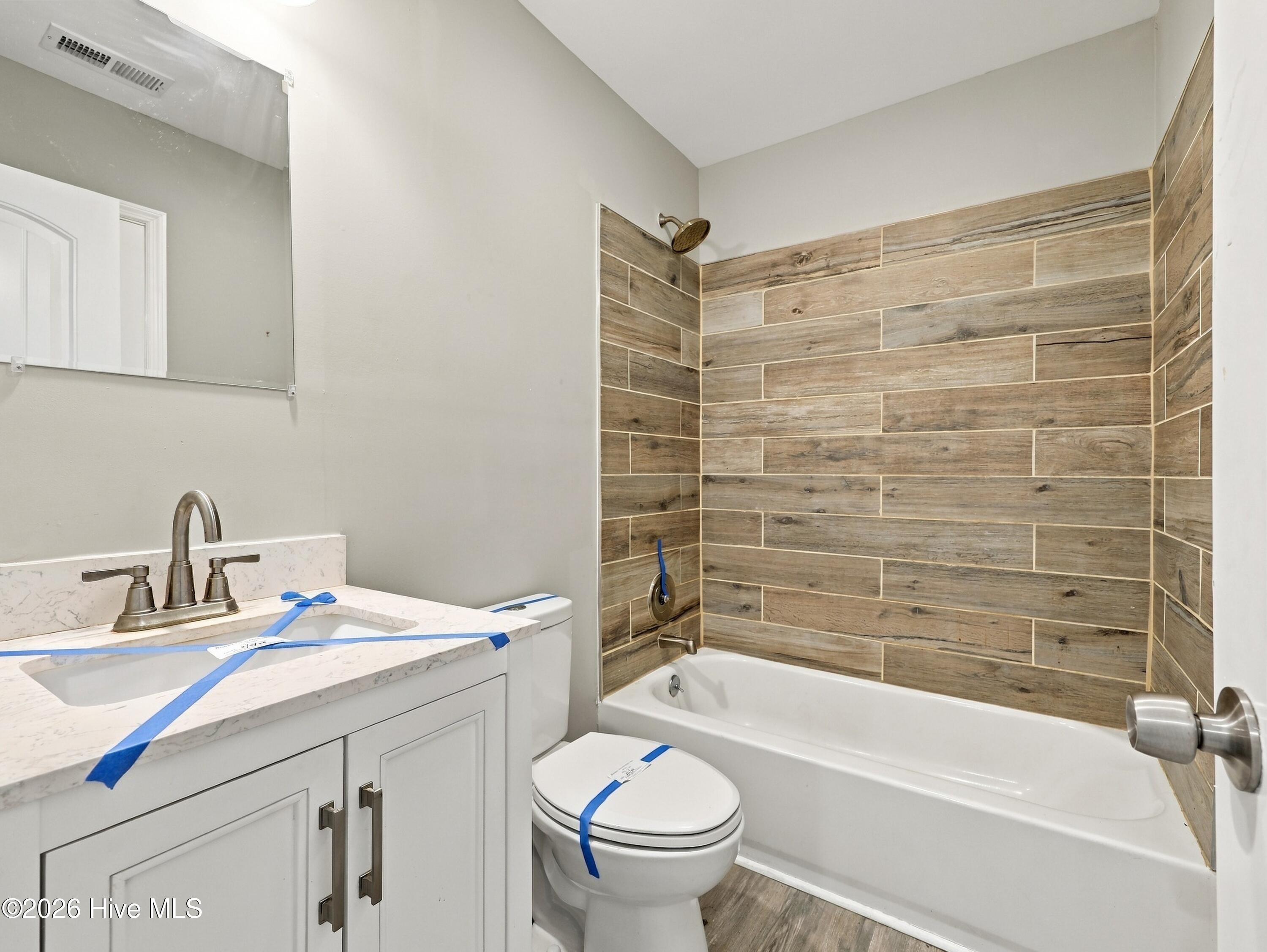 Bathroom with a white vanity and bathtub surrounded by wood-look tile; blue protective tape remains on fixtures.