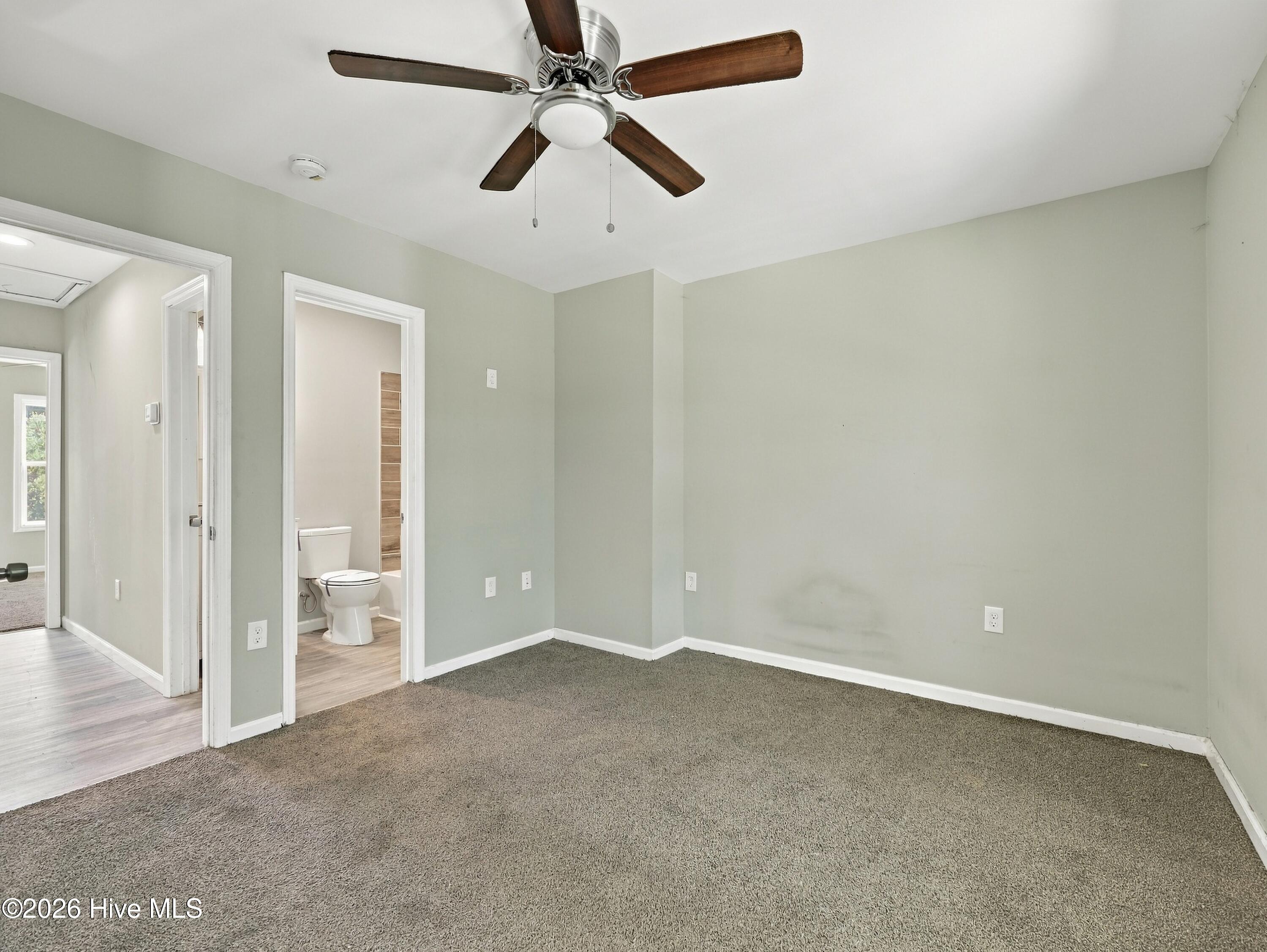 Empty bedroom with gray carpet and ceiling fan, opening to a bathroom.