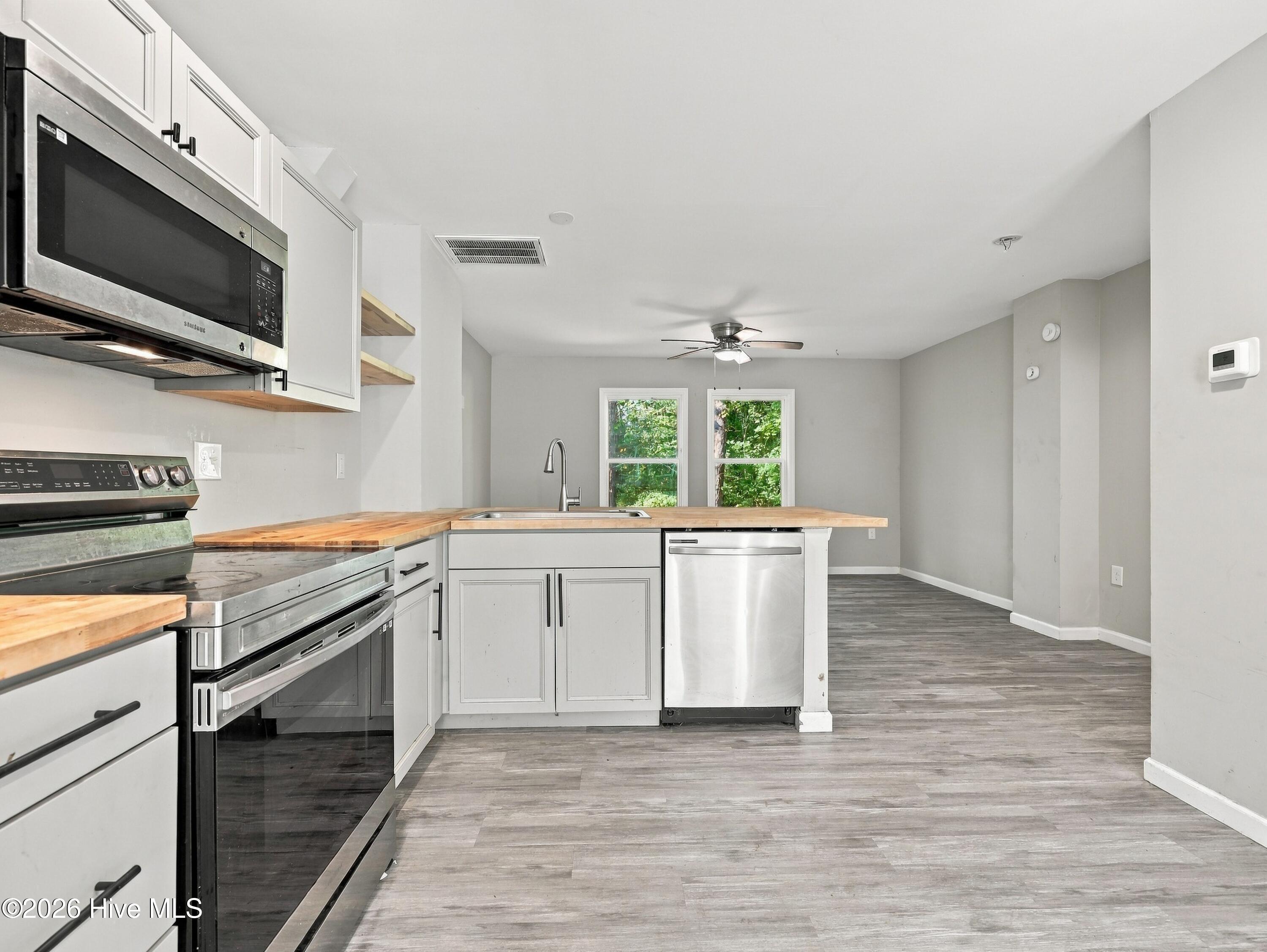 Kitchen with butcher-block counters and stainless appliances, opening into an empty room with gray plank floors.