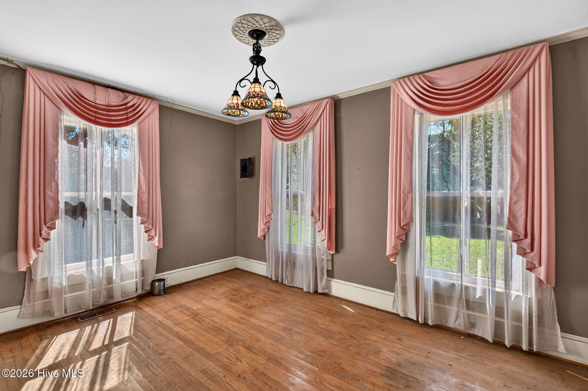 Empty room with hardwood floors, three tall windows with pink drapes, and a decorative chandelier.