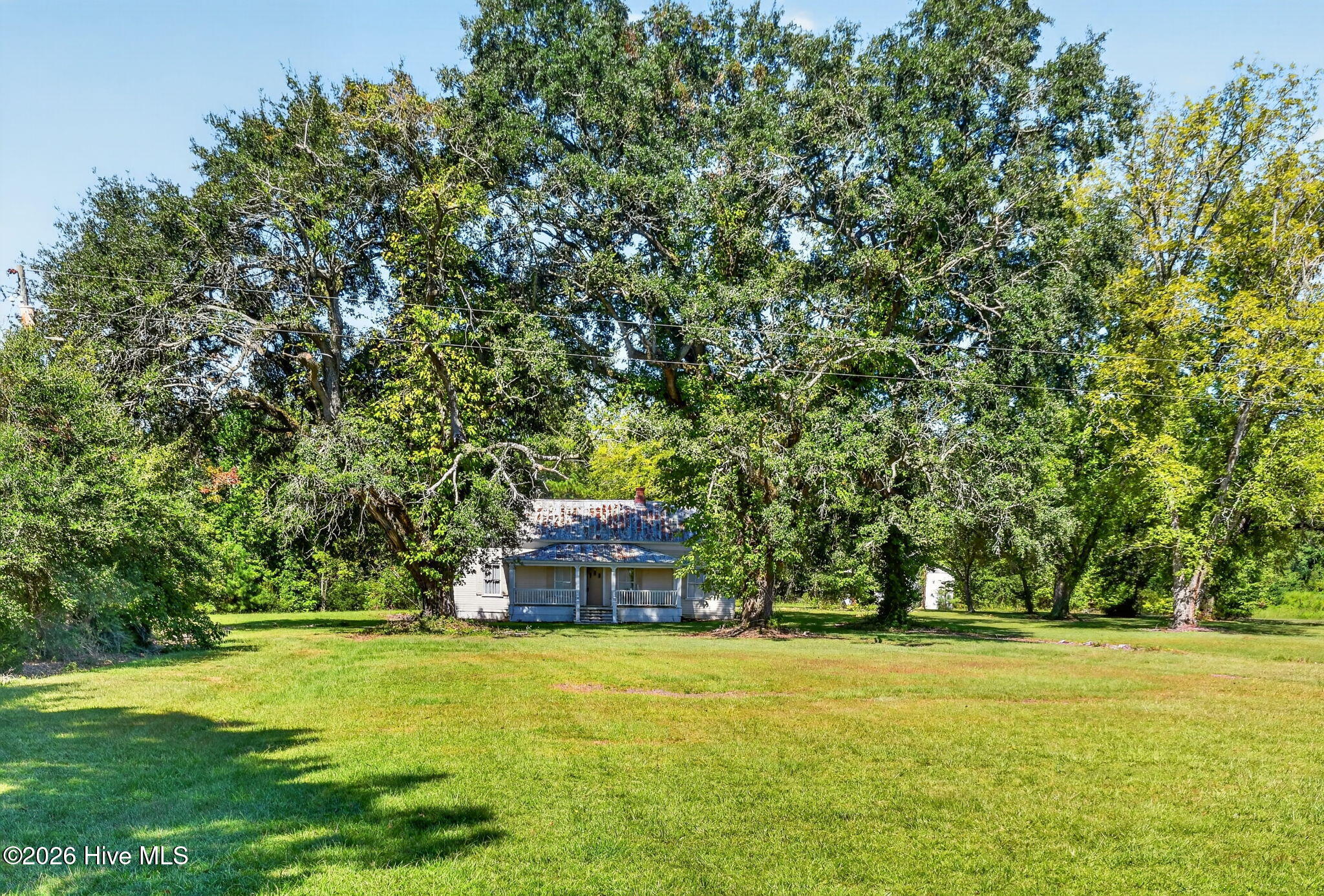 Small house with a covered porch beneath mature trees, set behind a broad lawn.