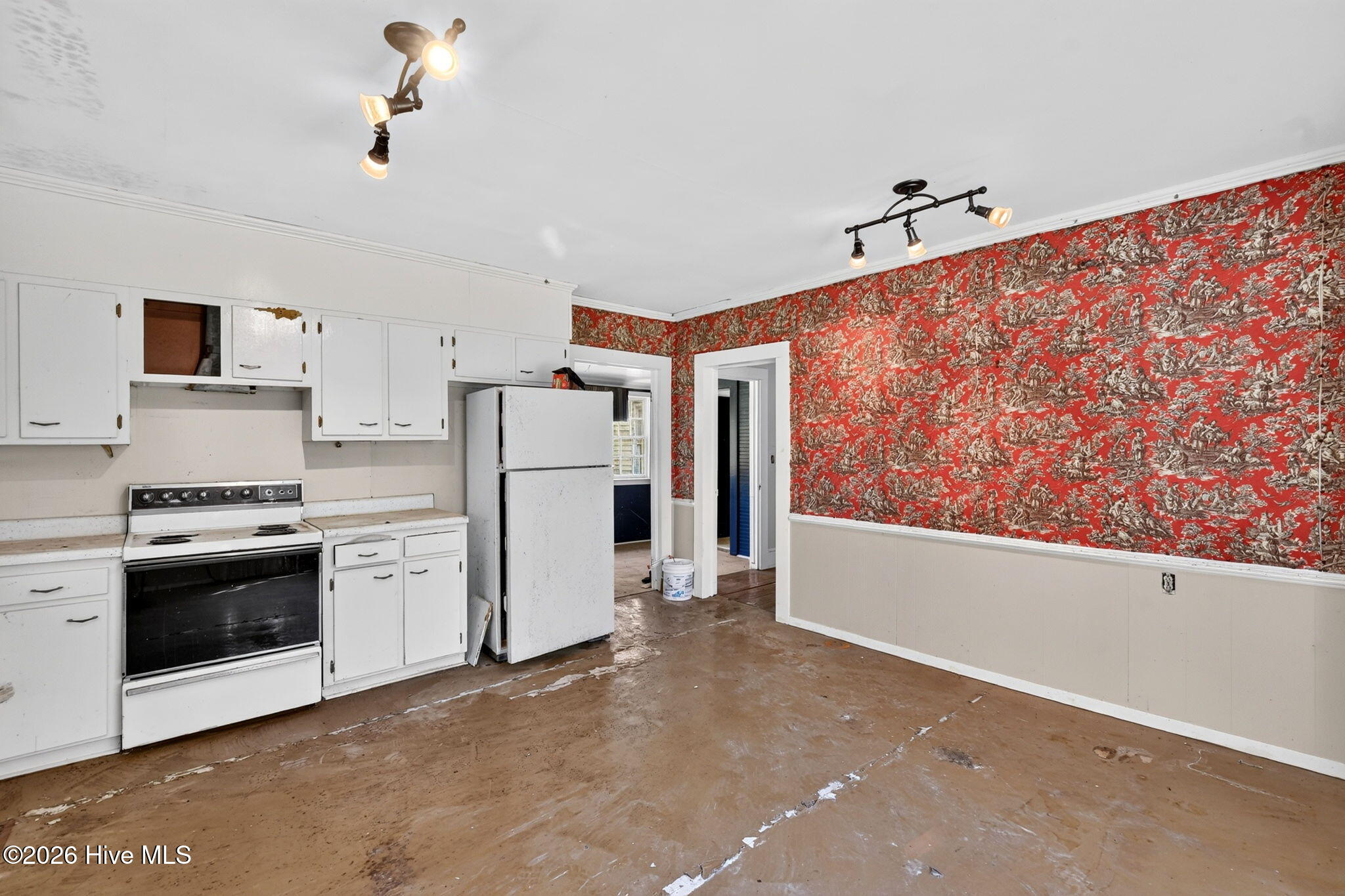 Vacant kitchen with red patterned wallpaper, white cabinets, and worn wood flooring.