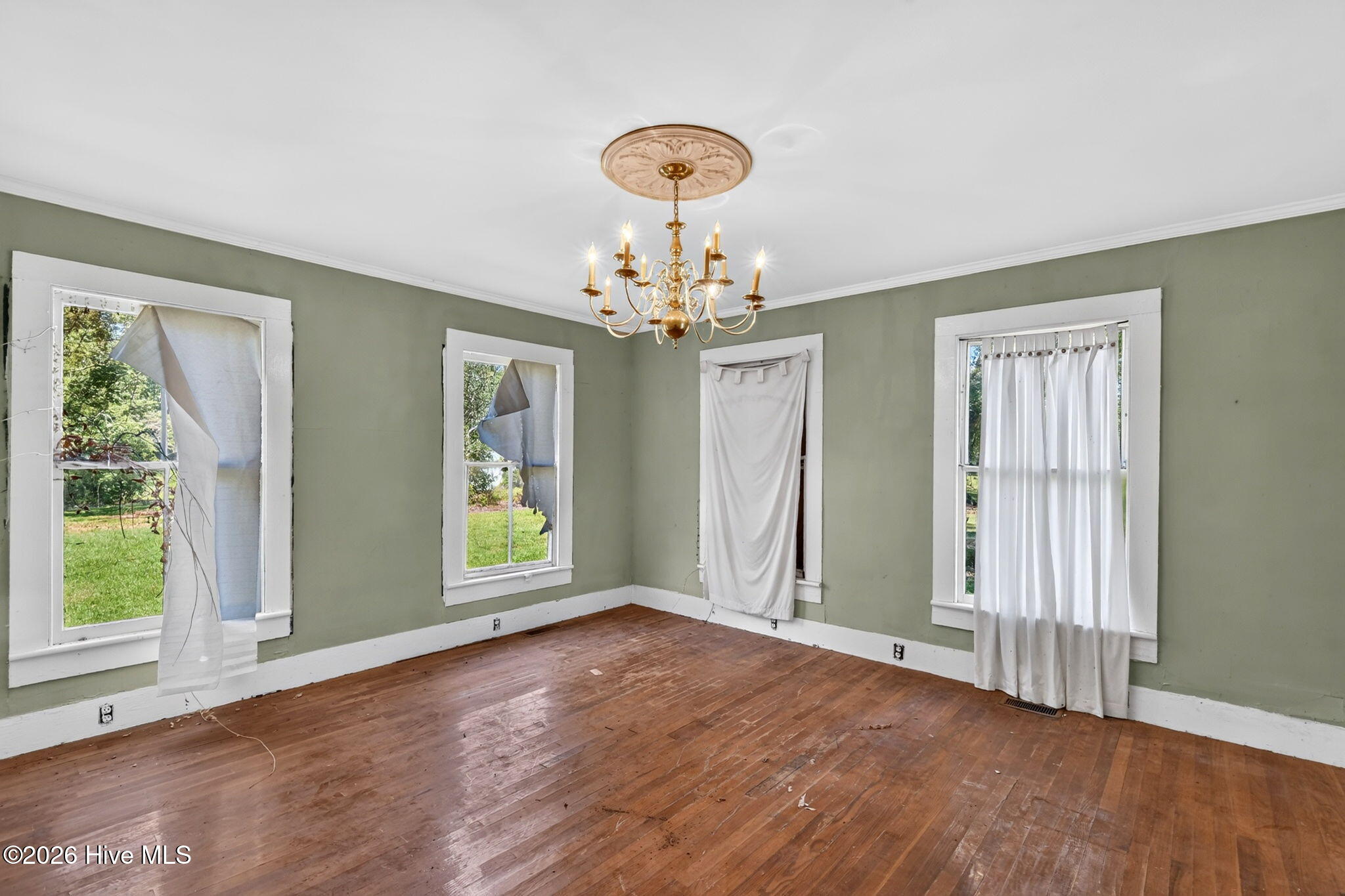 Empty room with green walls, worn hardwood floors, curtained windows, and an ornate brass chandelier.