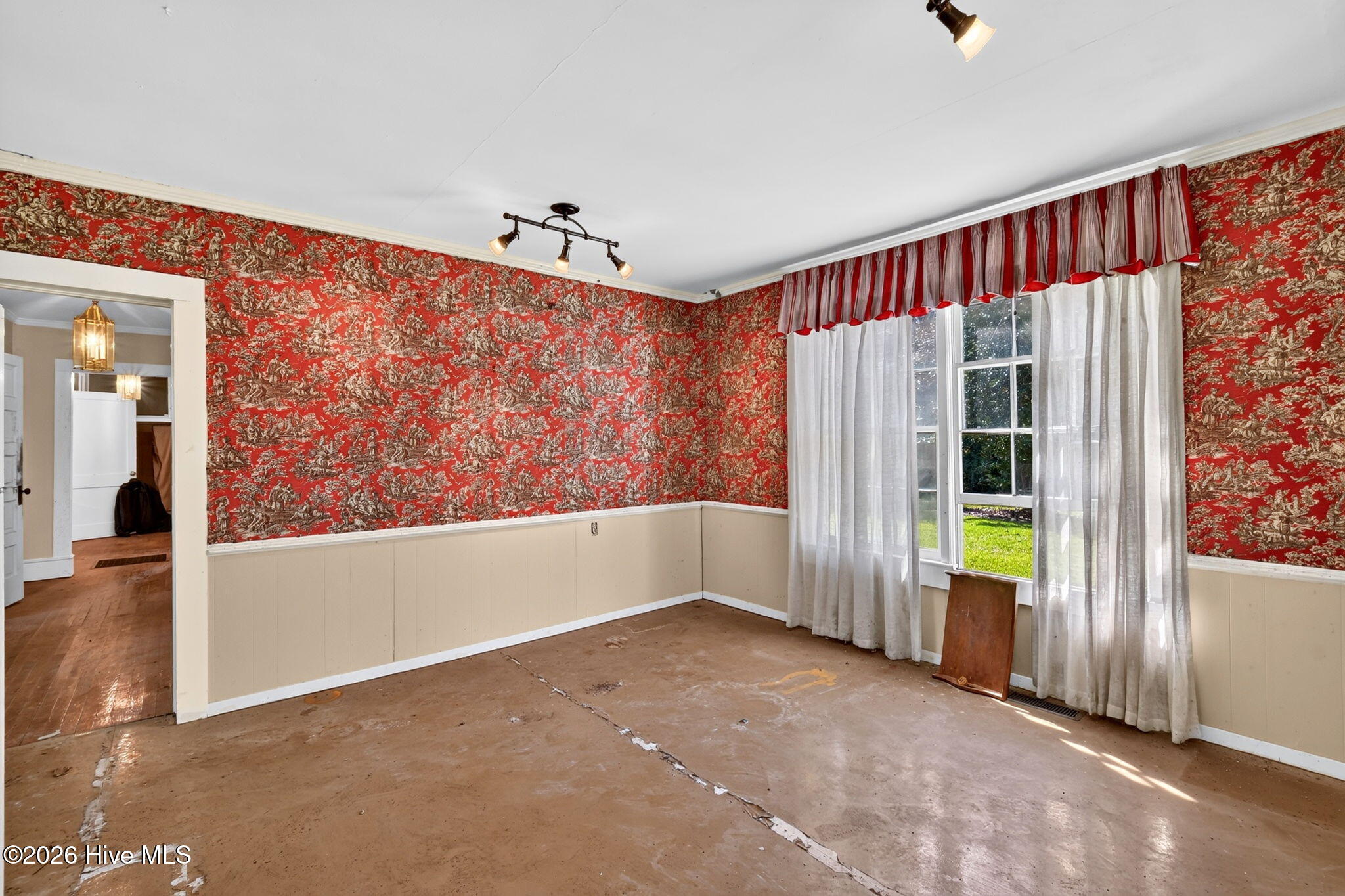 Empty room with red patterned wallpaper, beige wainscoting, a curtained window, and a worn floor.