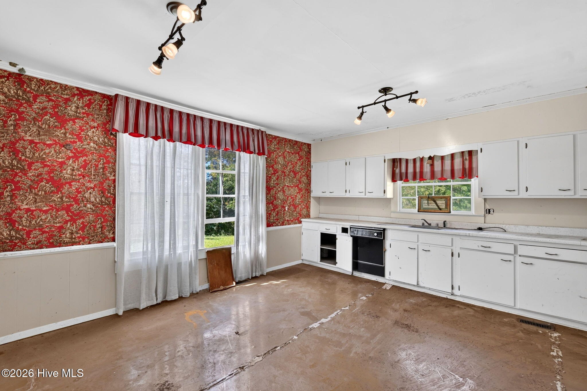 Kitchen with white cabinets, red patterned wallpaper, sheer curtains, and a worn floor.