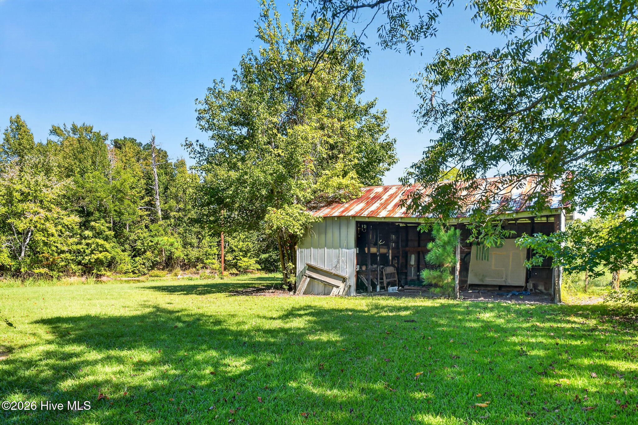 Open-sided outbuilding with a rusted metal roof beside a grassy lawn and wooded yard.