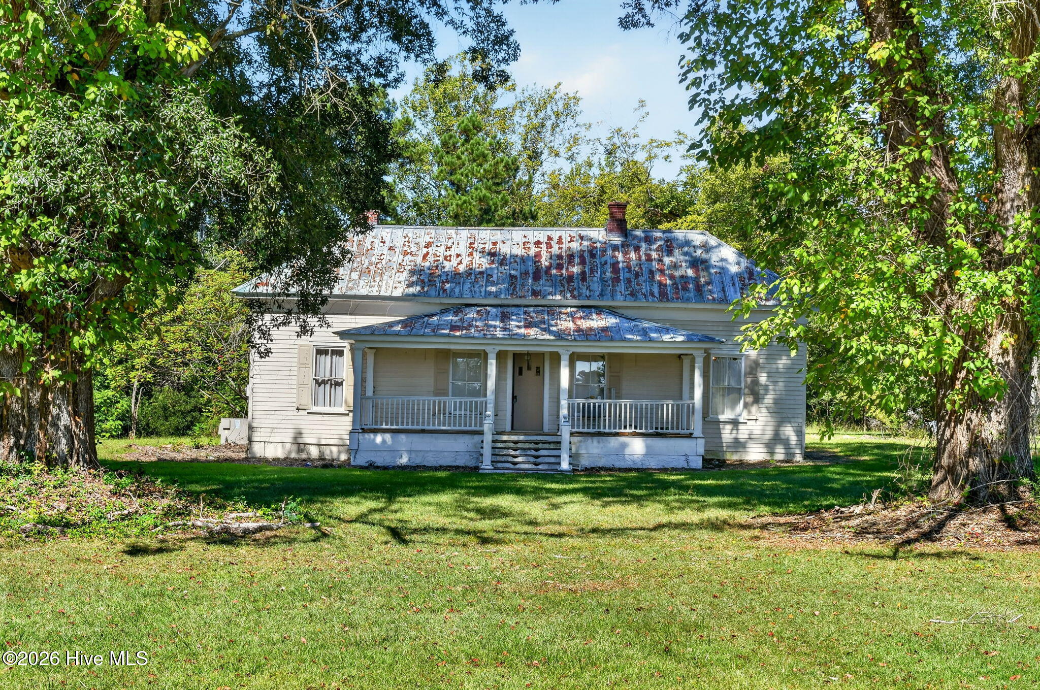 Light-colored house with a covered porch and weathered metal roof, framed by mature trees.
