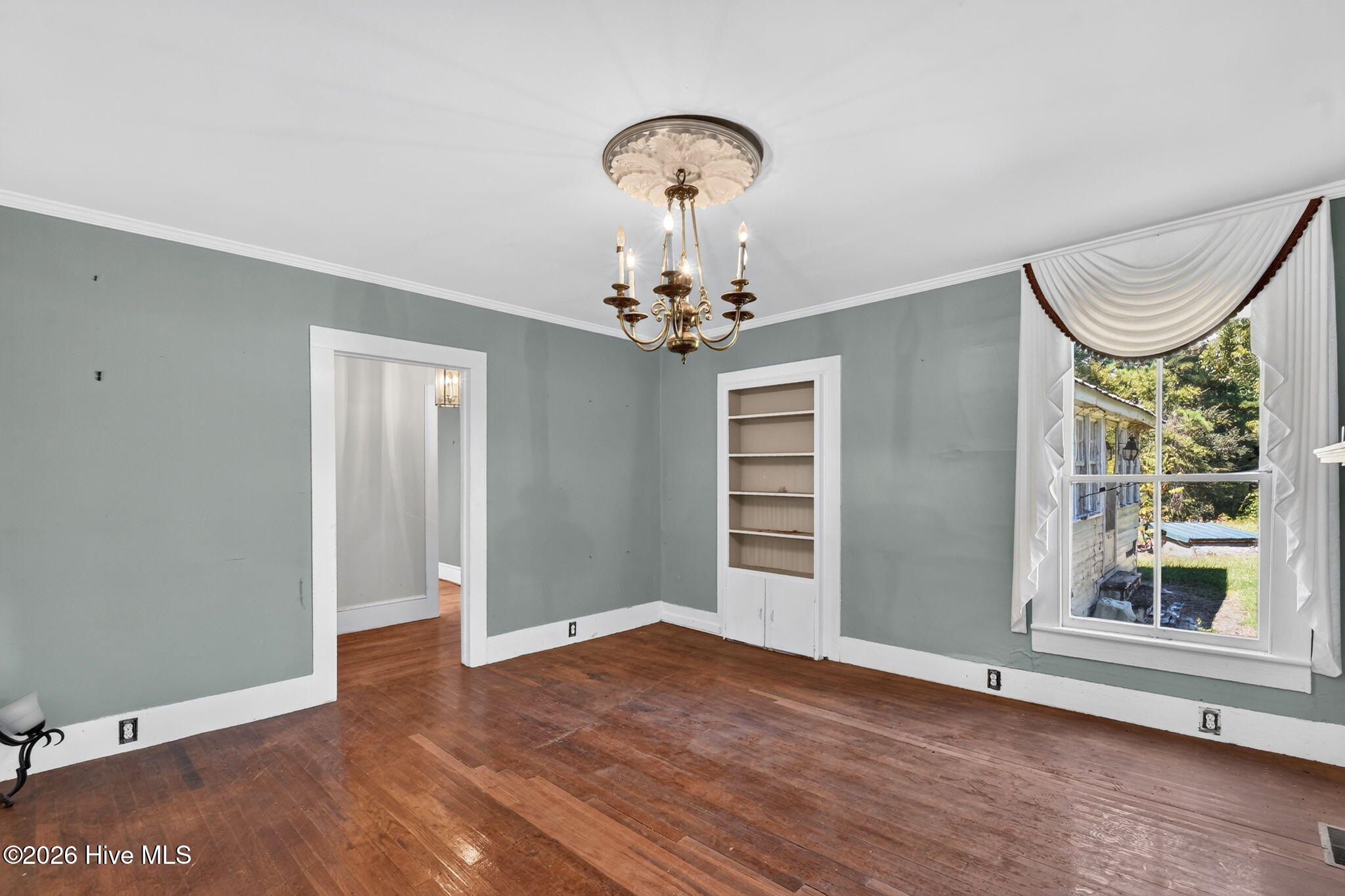 Vacant room with hardwood floors, a brass chandelier, built-in shelves, and a large curtained window.