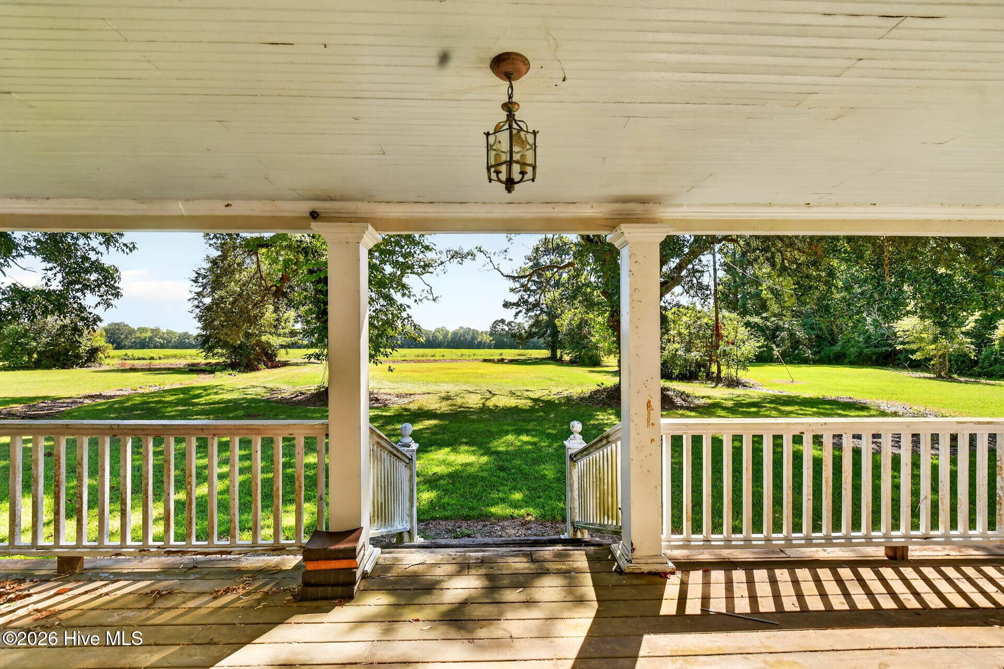Covered porch with white railings and columns overlooking a sunny lawn and mature trees.