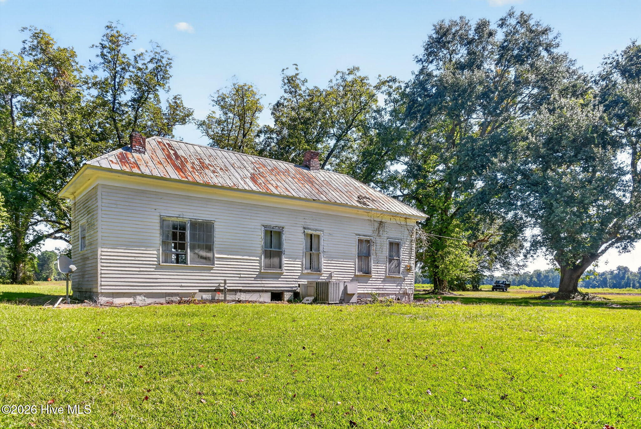 White single-story house with a weathered metal roof and mature trees surrounding the grassy lawn.