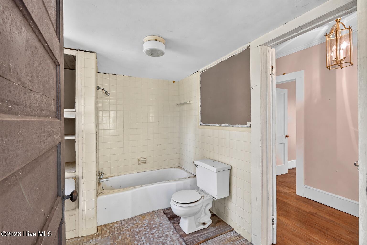Older tiled bathroom with a bathtub and toilet, opening onto a pink room with hardwood floors.
