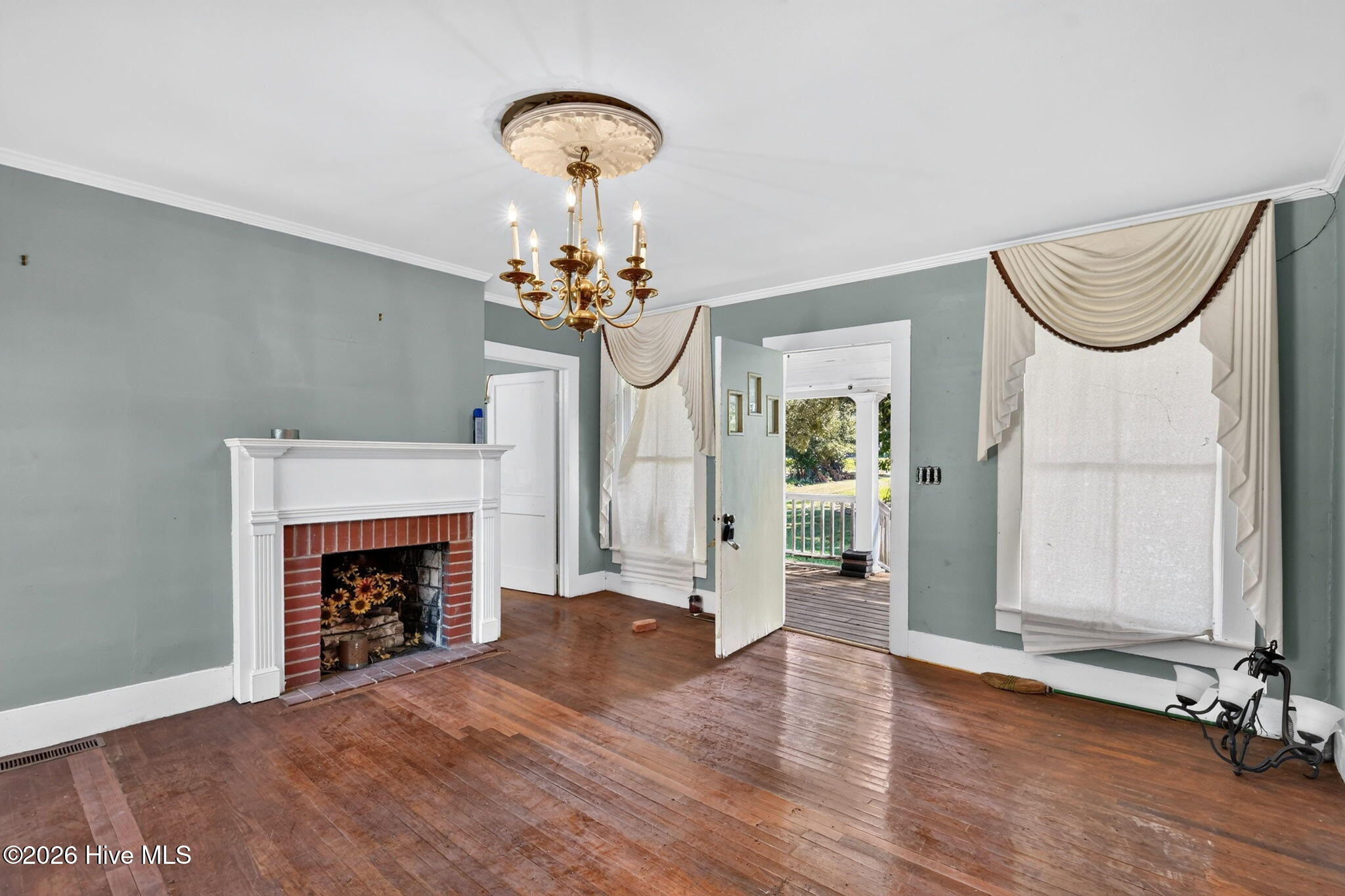 Vacant living room with a brick fireplace, hardwood floors, brass chandelier, and open doorway to a porch.