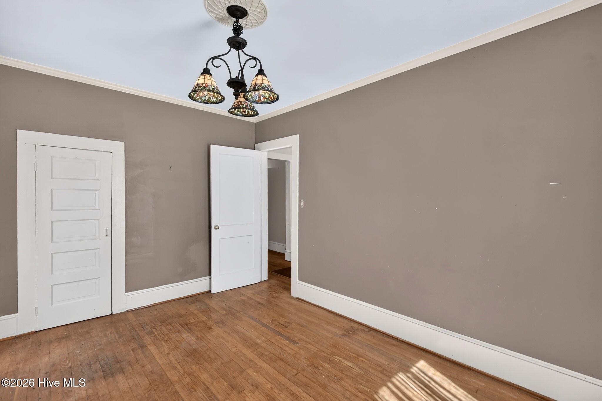 Empty room with hardwood floors, taupe walls, white doors, and a decorative chandelier.