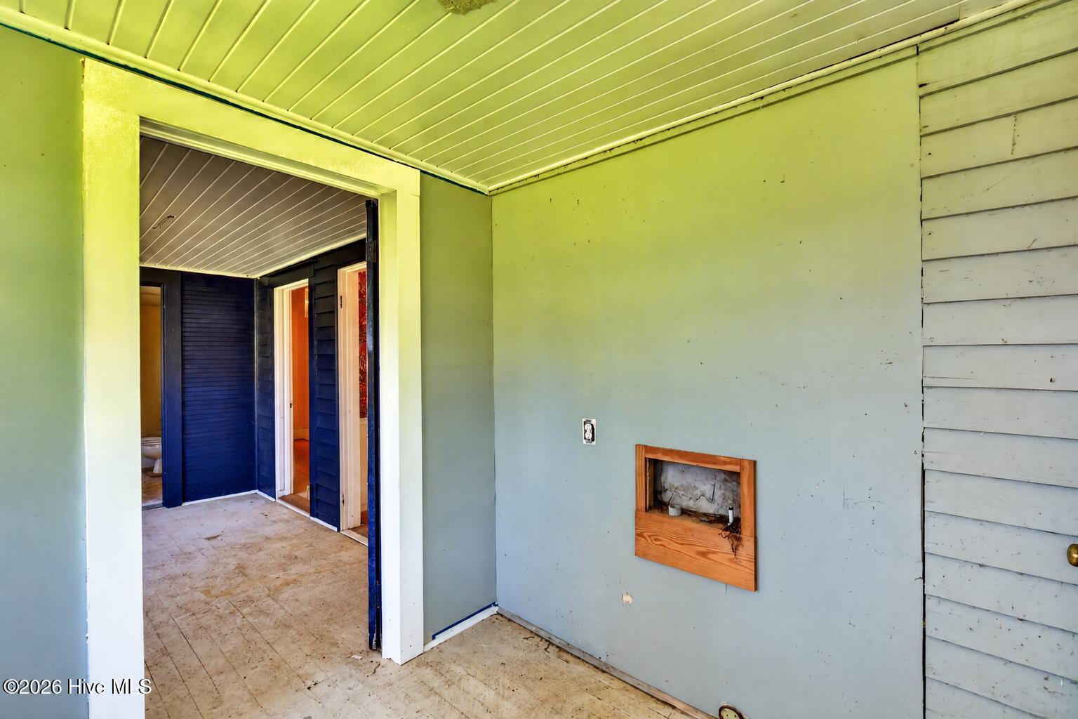 Vacant unfinished room with pale green walls, paneled ceiling, and doorway to a dark blue hallway.