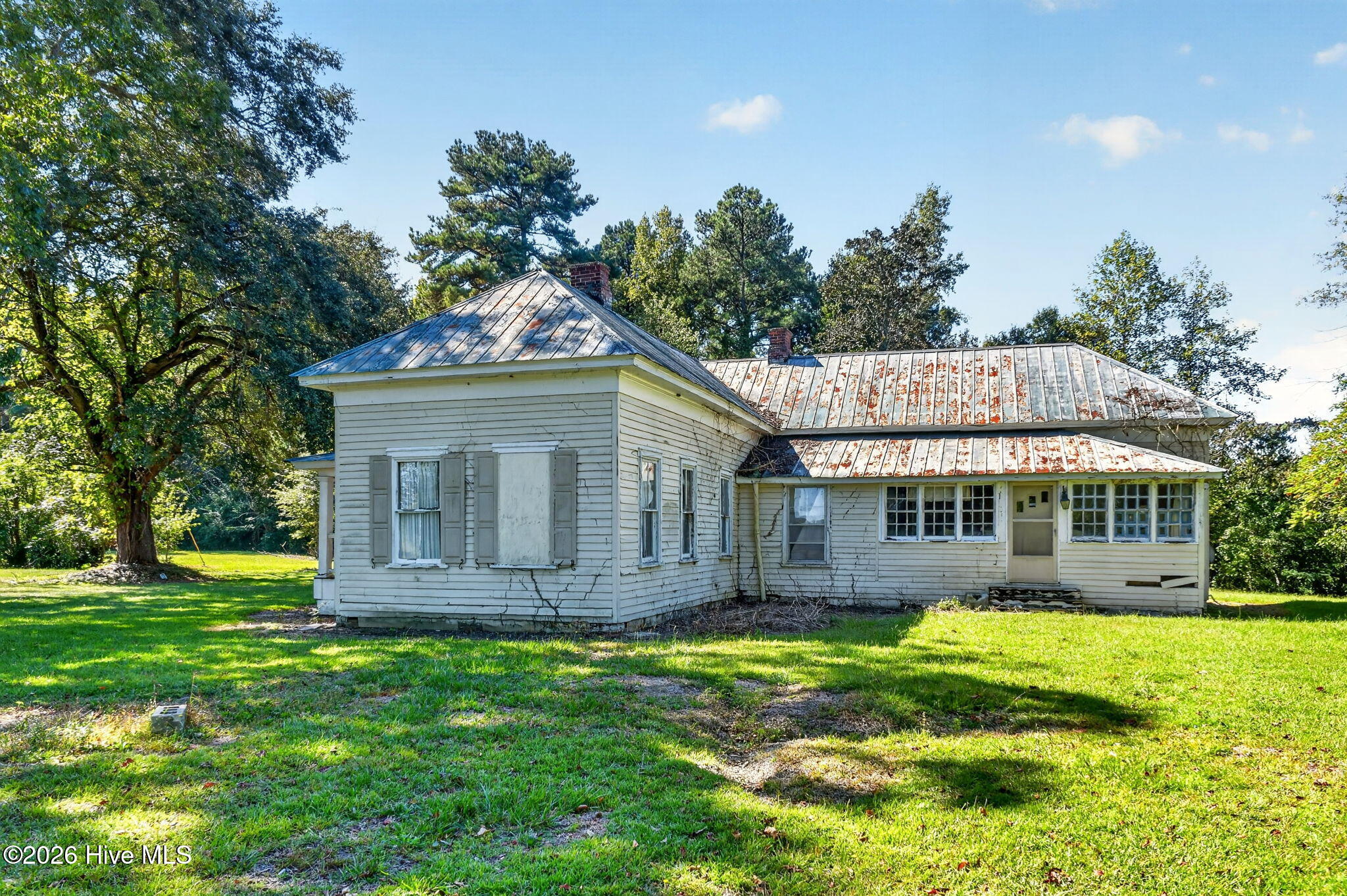 White house with a rust-spotted metal roof on a grassy lawn beneath mature trees.