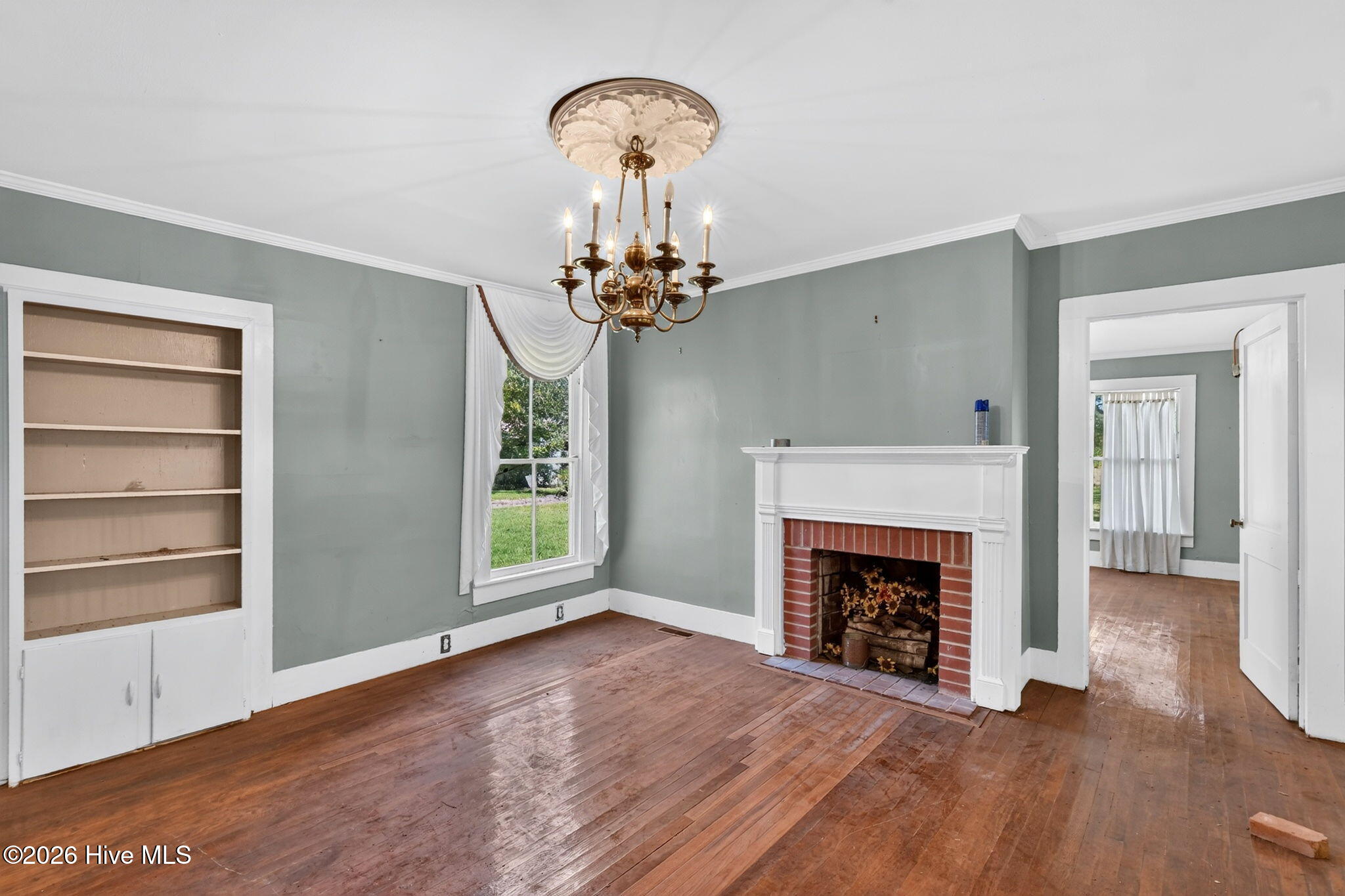 Empty living room with a brick fireplace, built-in shelves, hardwood floors, and brass chandelier.