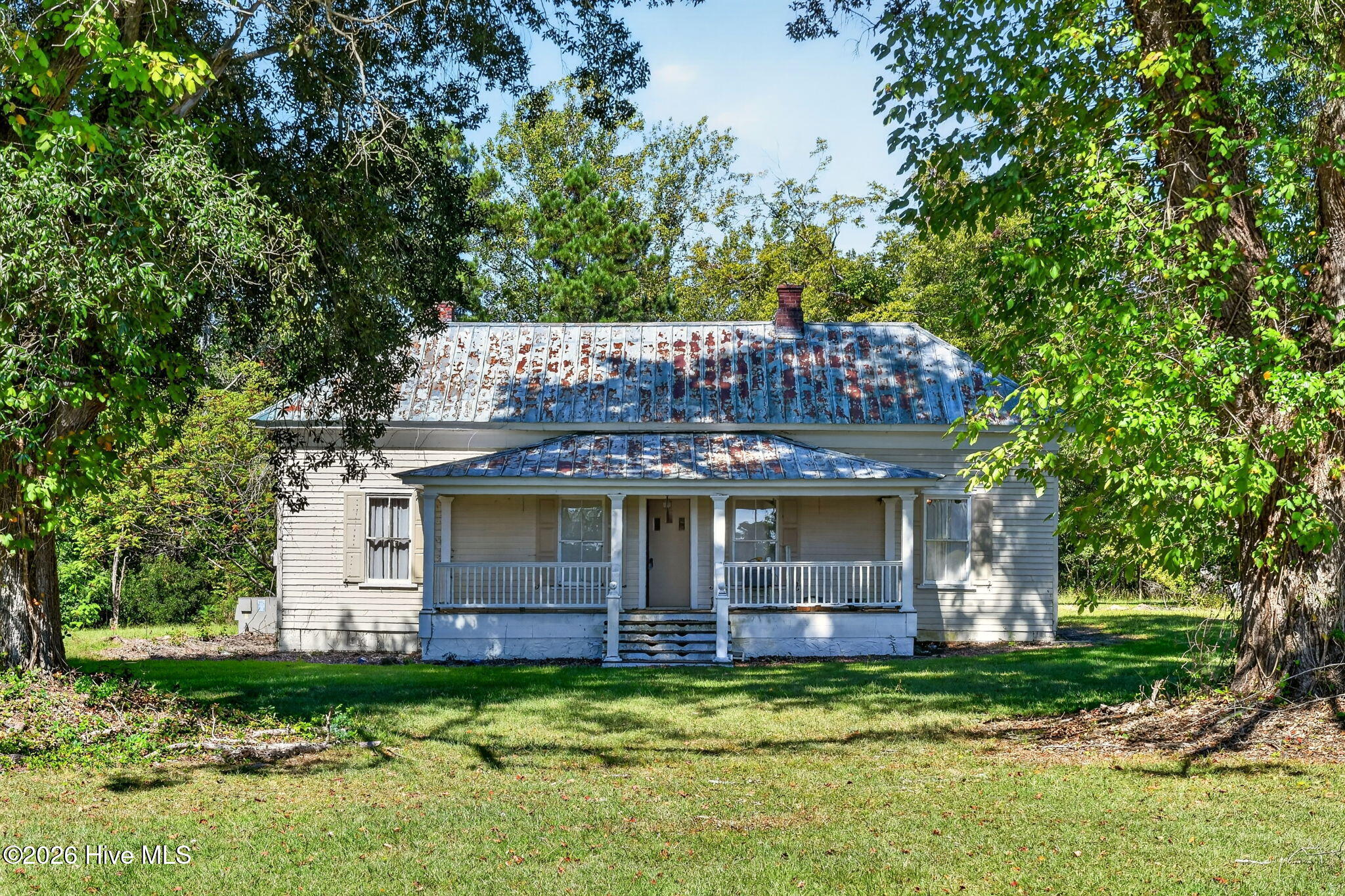 Single-story house with a covered front porch and weathered metal roof beneath mature trees.