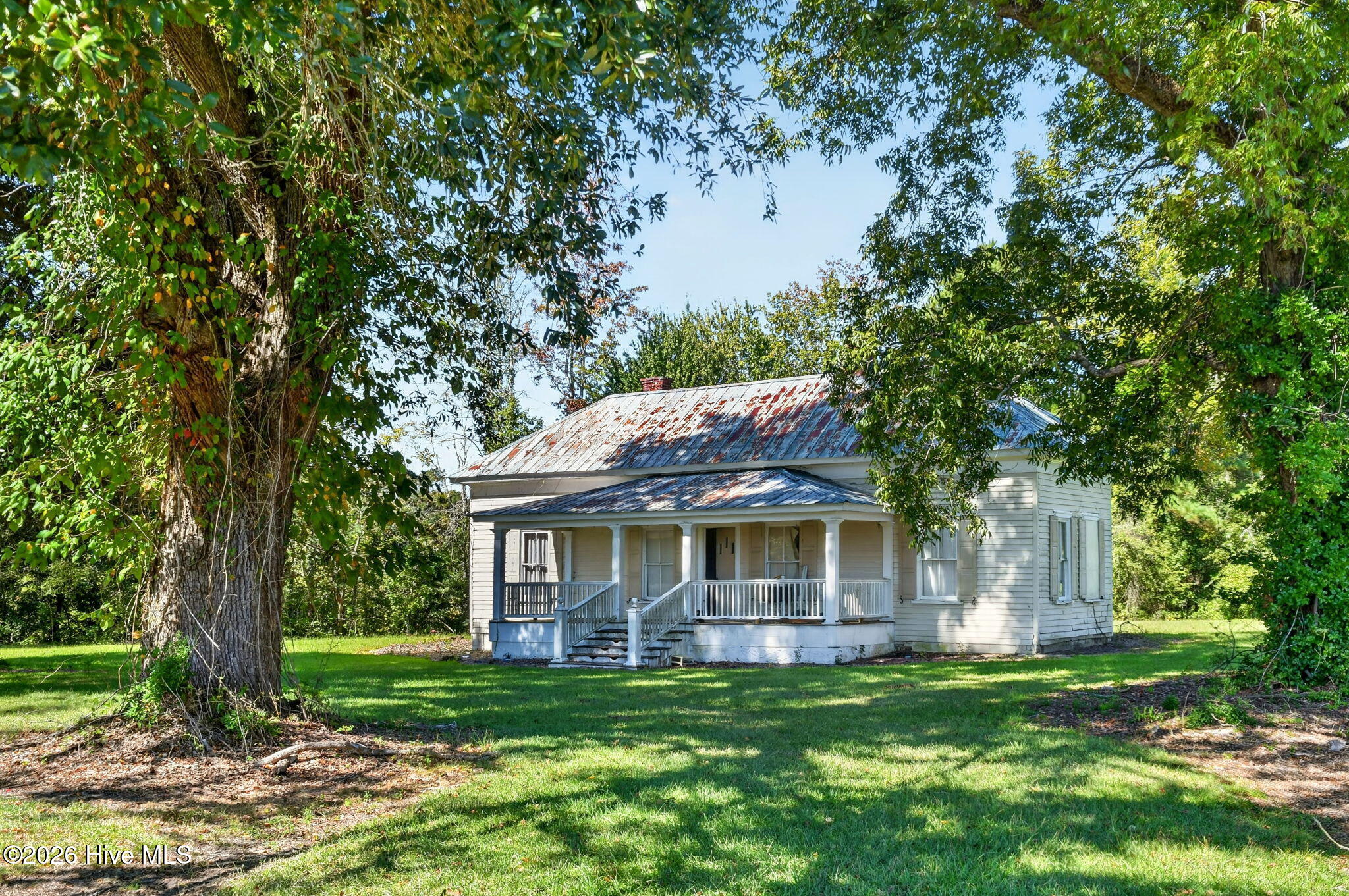White-sided house with a weathered metal roof and covered porch beneath mature trees.