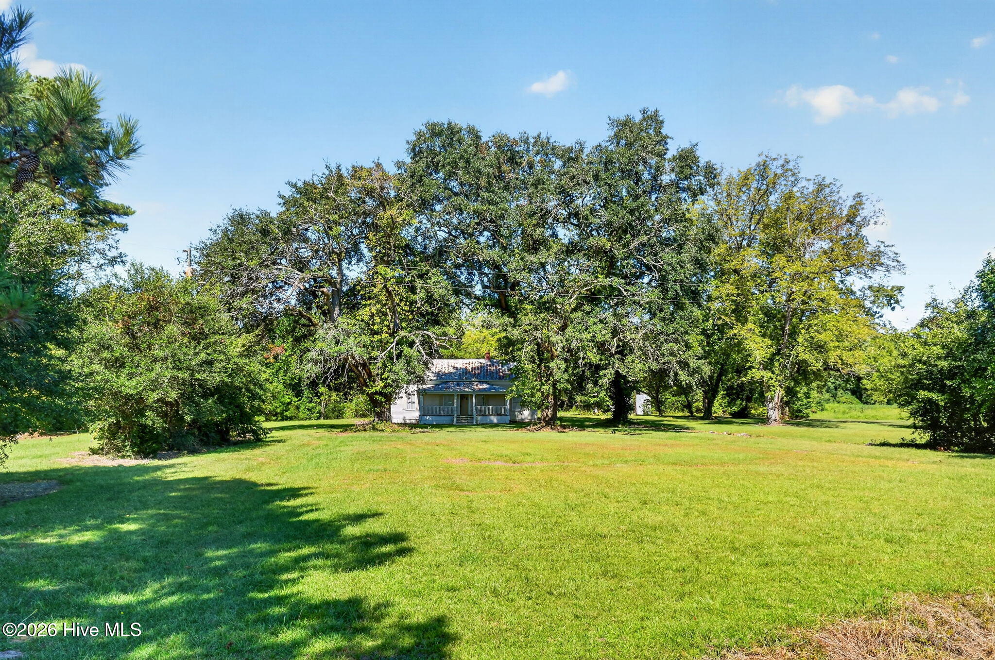 Small white house with a covered porch beneath mature trees, beyond a broad green lawn.