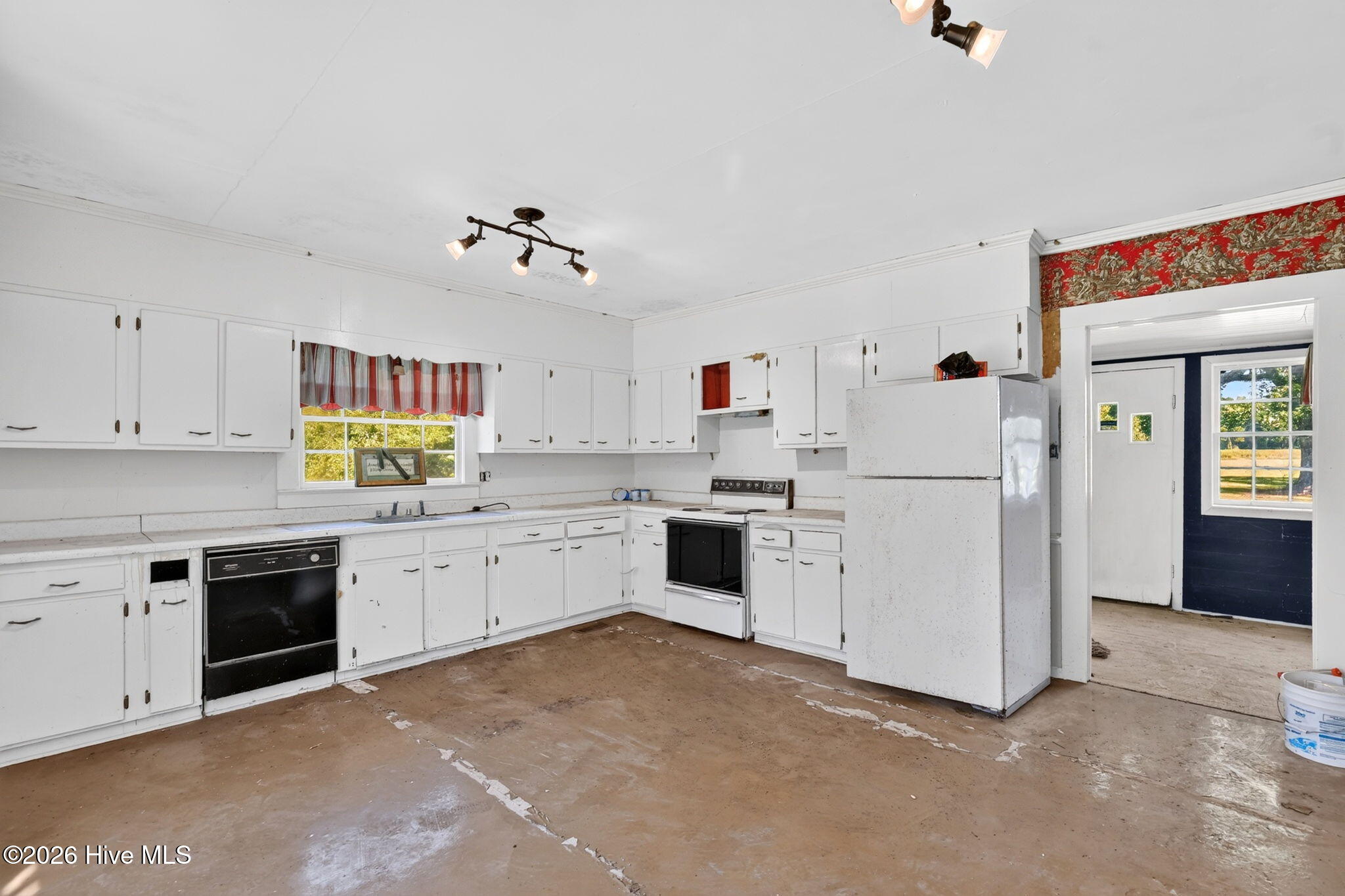 Vacant kitchen with white cabinets, older appliances, red-striped window curtains, and worn brown flooring.