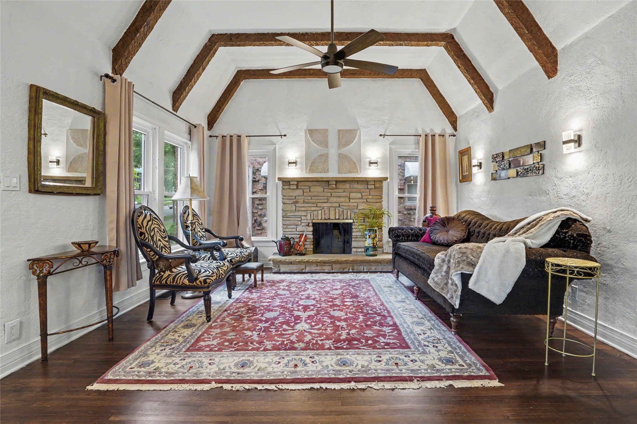 Living room with exposed wood ceiling beams, a stone fireplace, and a large patterned rug.