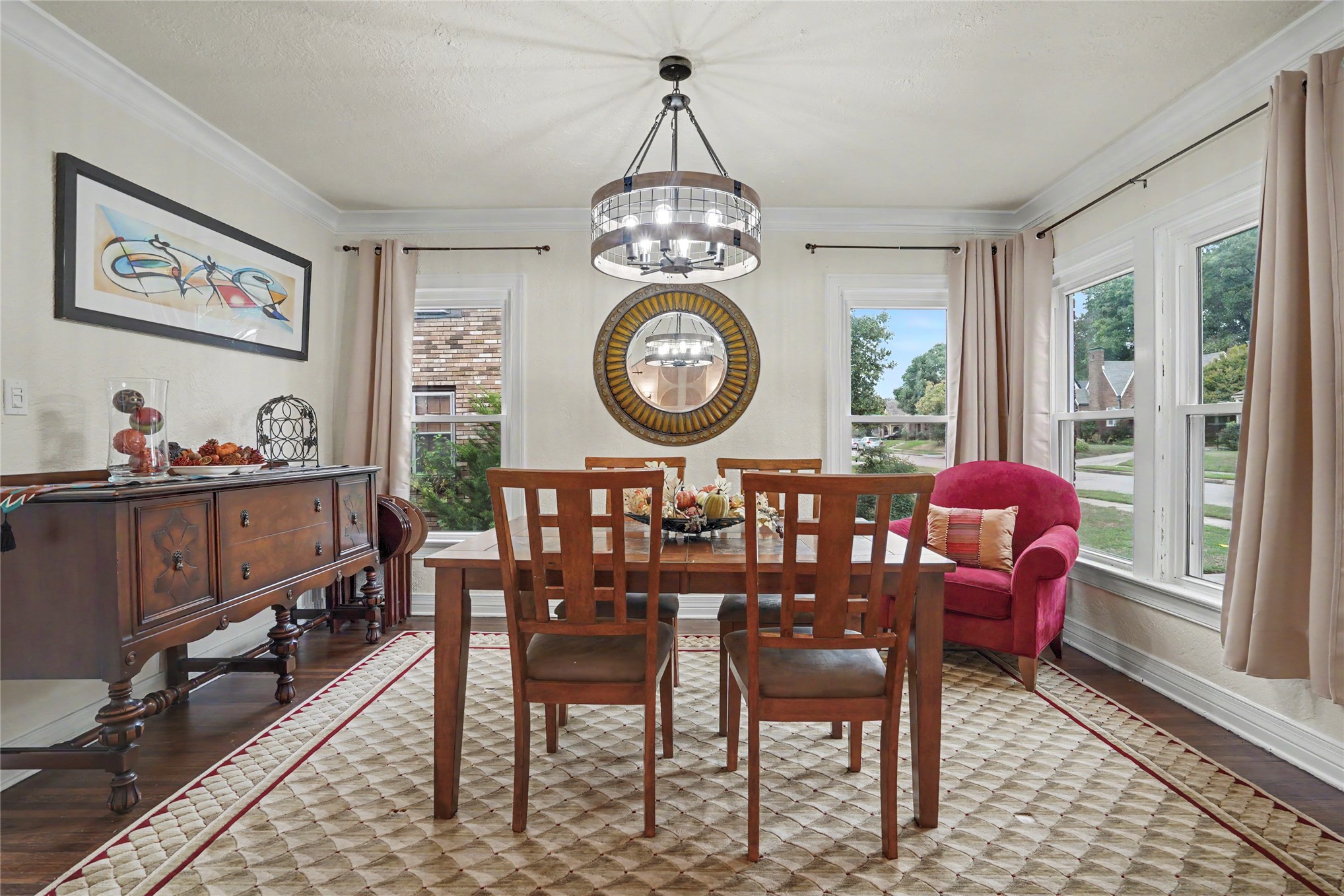 Dining room with a wooden table, circular chandelier, large windows, and bright pink armchair.