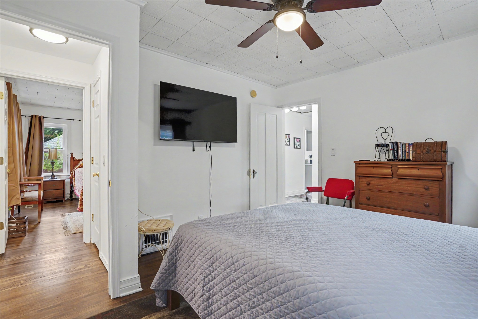 Bedroom with a gray quilted bed, wall-mounted TV, ceiling fan, and wooden dresser.