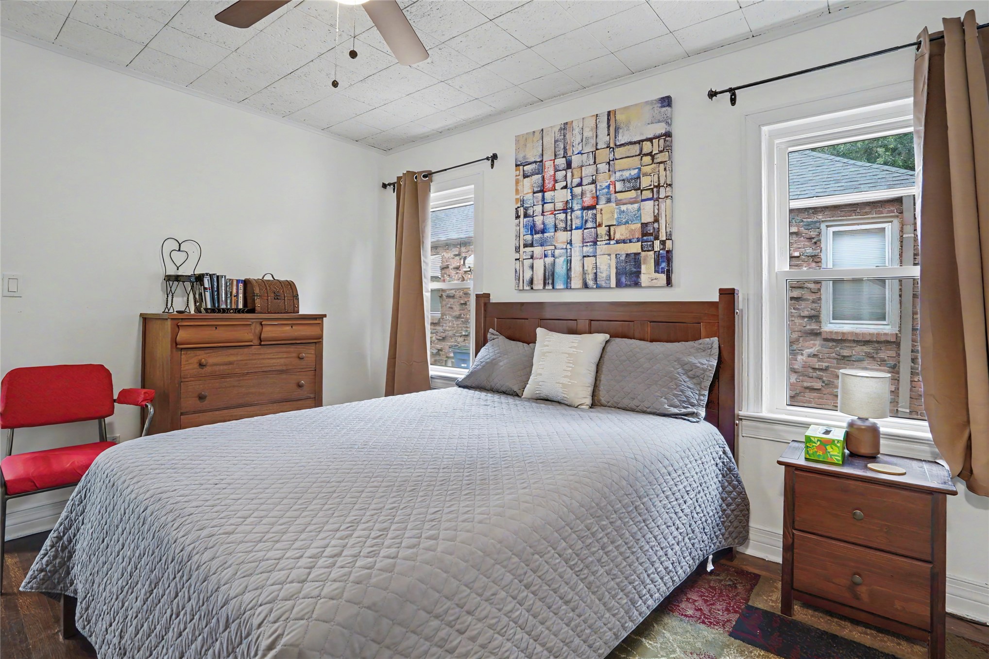 Bedroom with a gray quilted bed, wooden furniture, abstract wall art, and two windows.