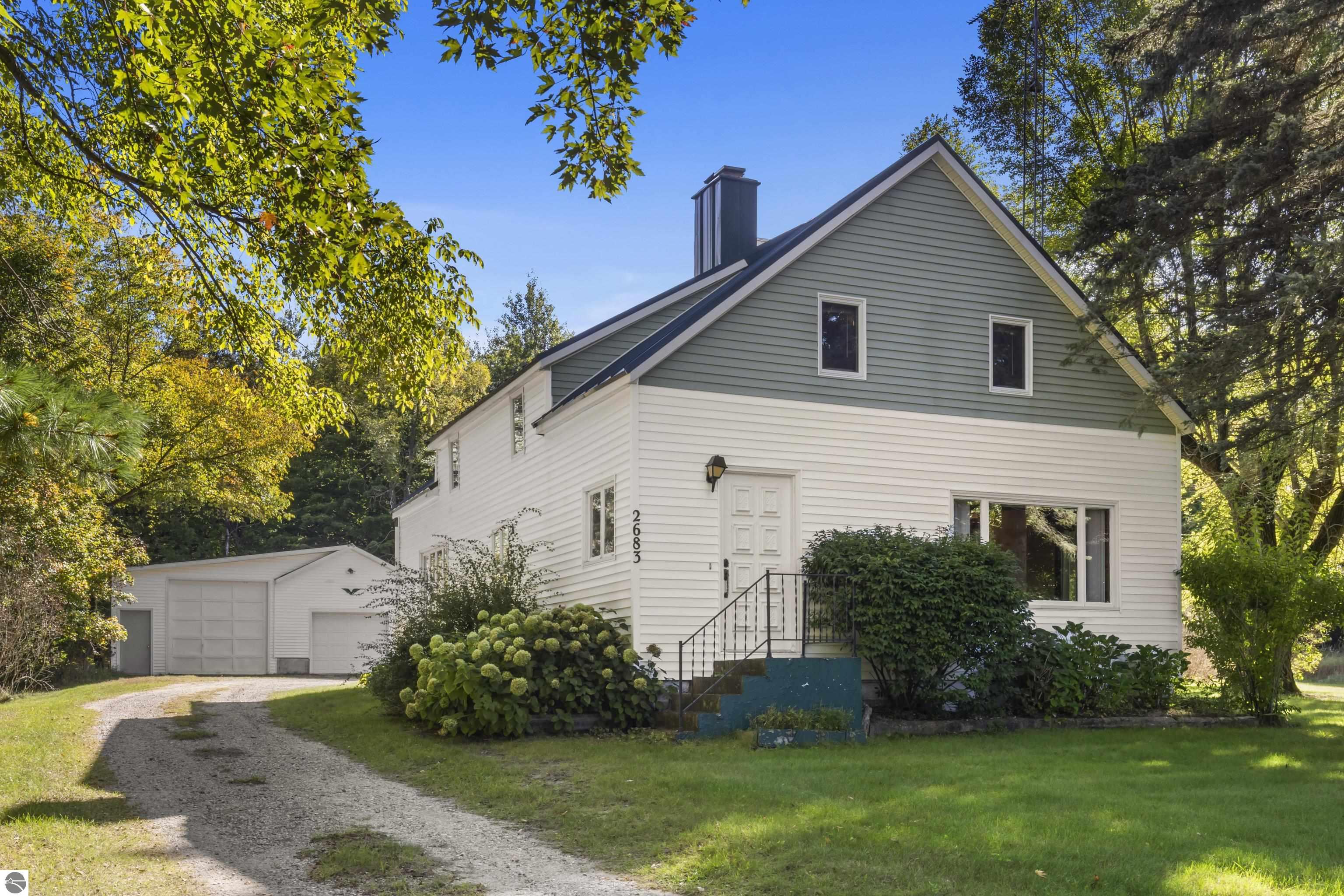 White two-story house with gray-green gable, gravel driveway, and detached garage among autumn trees.