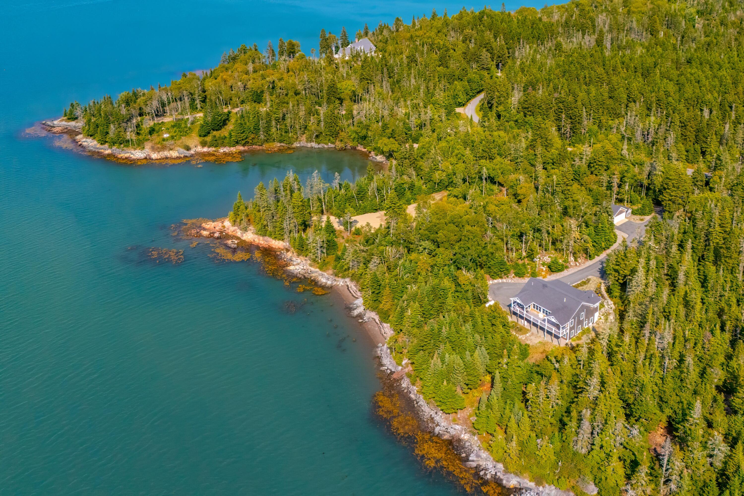 Aerial view of a gray coastal home beside a rocky, forested shoreline.