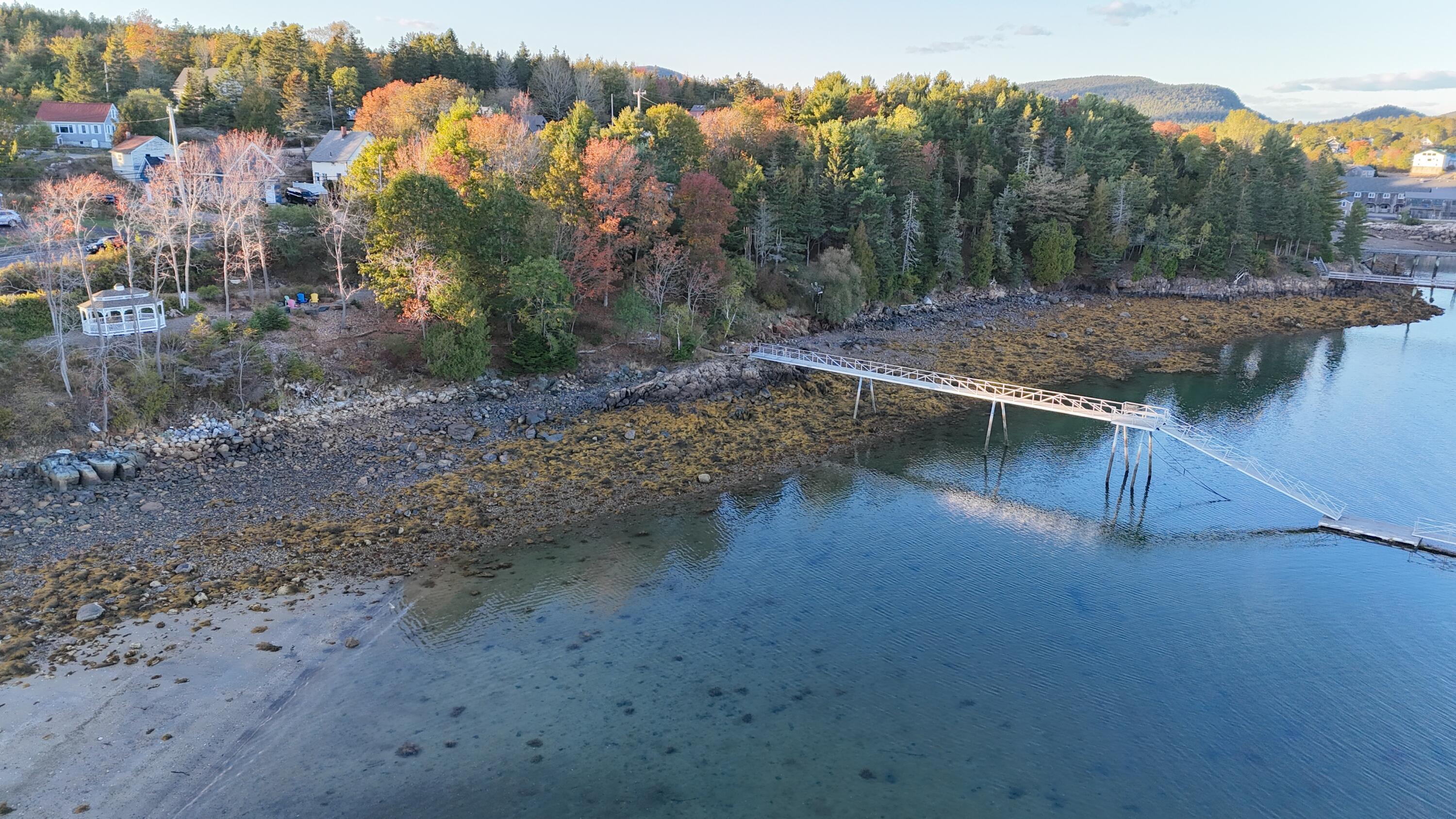 Aerial waterfront view with a long dock, rocky tidal shore, and autumn-colored trees.