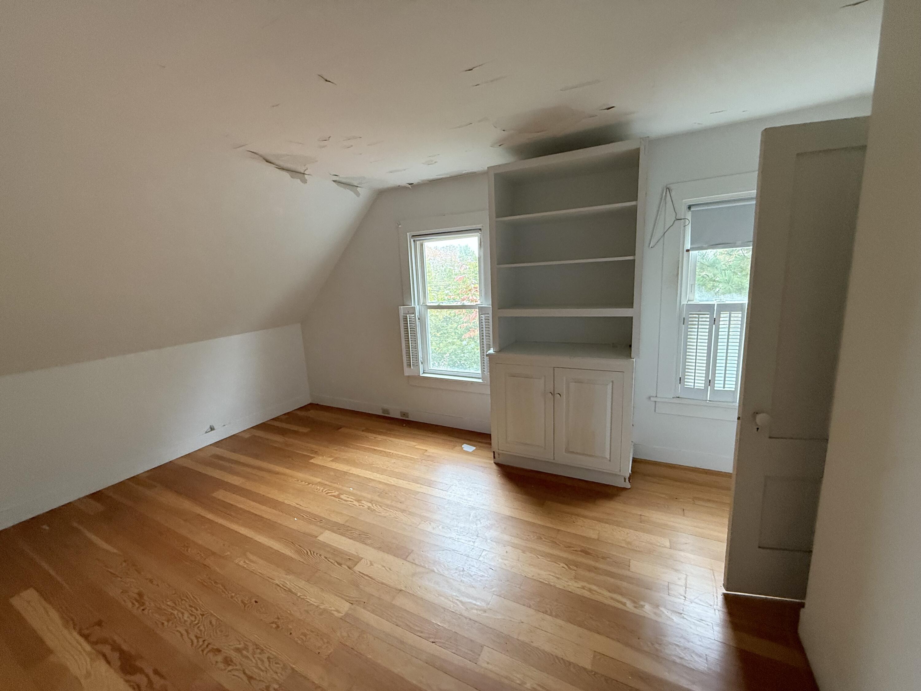 Empty attic bedroom with built-in shelving, hardwood floors, and peeling ceiling paint.