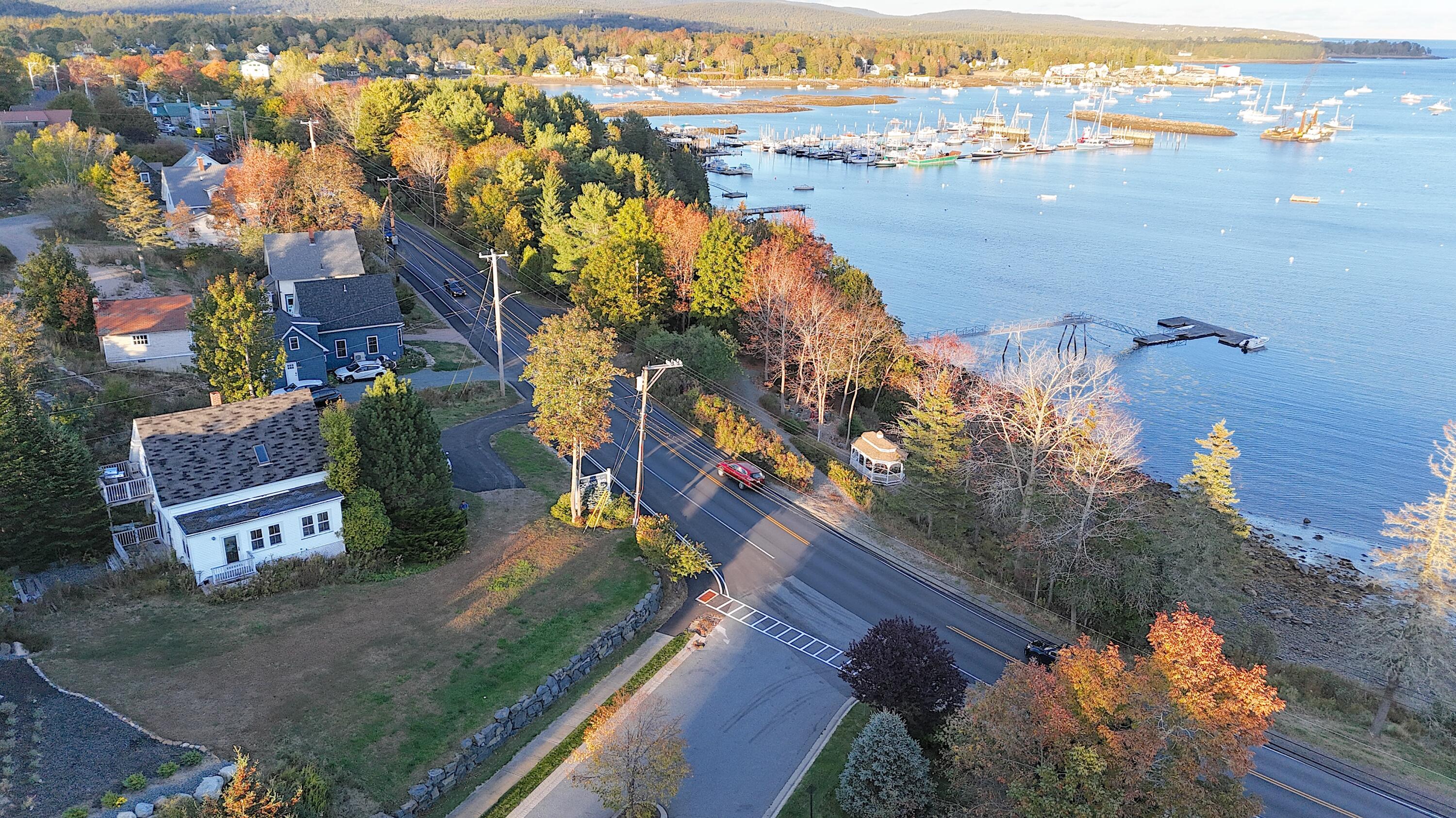 Aerial view of a coastal house beside the road, with autumn trees and a marina along the harbor.