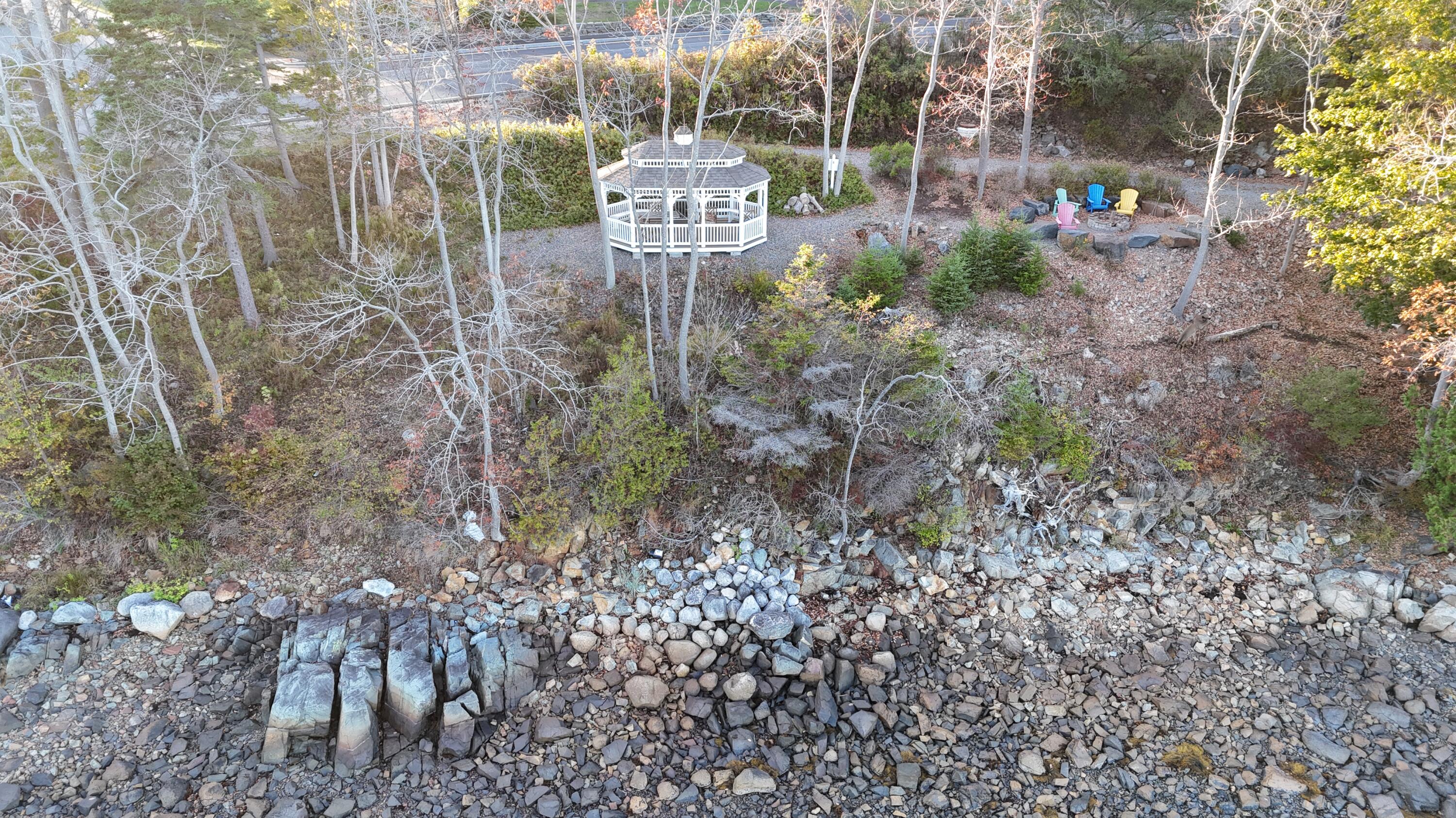 Aerial view of a rocky shoreline with a white gazebo and colorful chairs in a wooded yard.