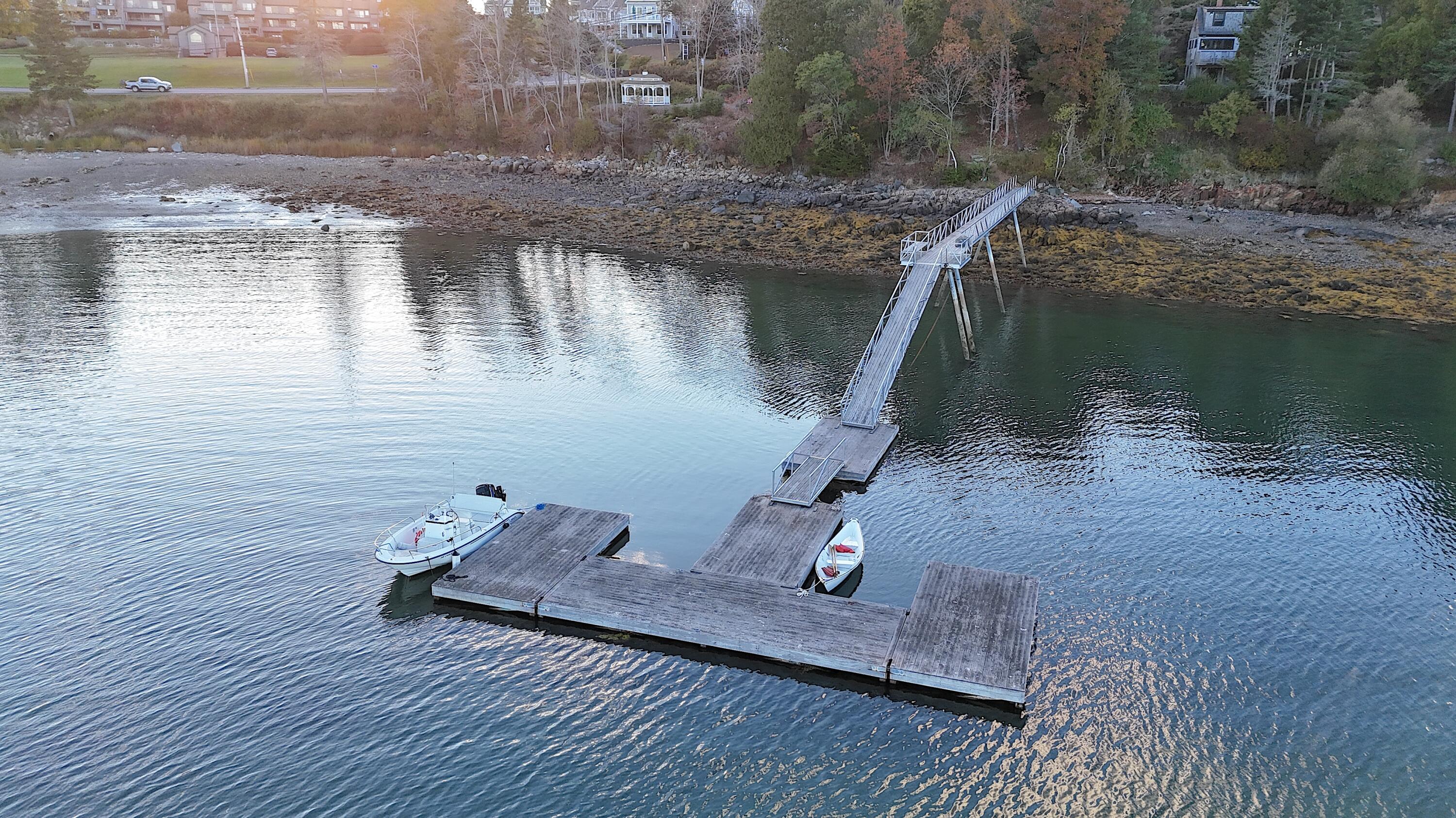 Waterfront dock with a long gangway, floating dock sections, and two boats beside a rocky shore.