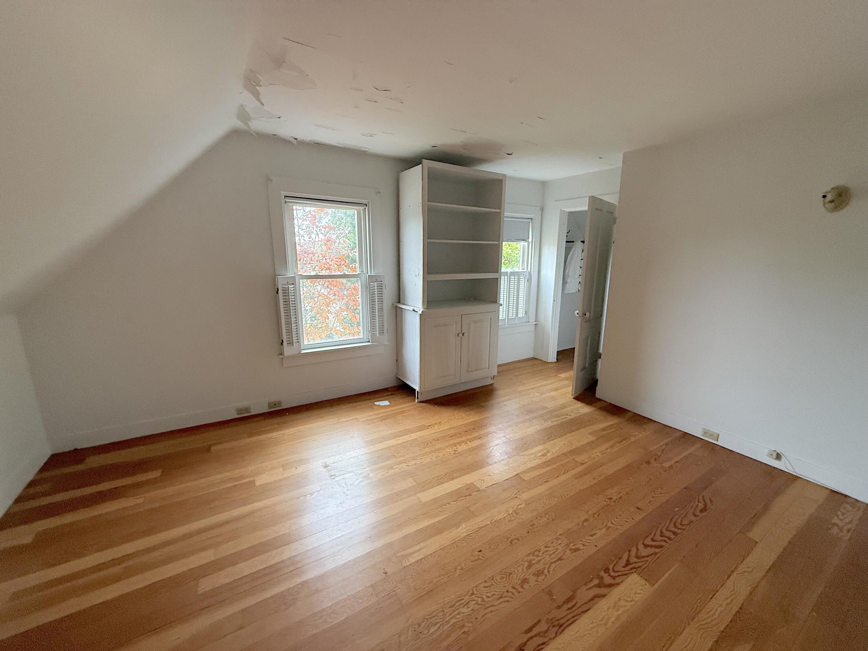Empty room with hardwood floors, built-in shelving, two windows, and peeling ceiling paint.
