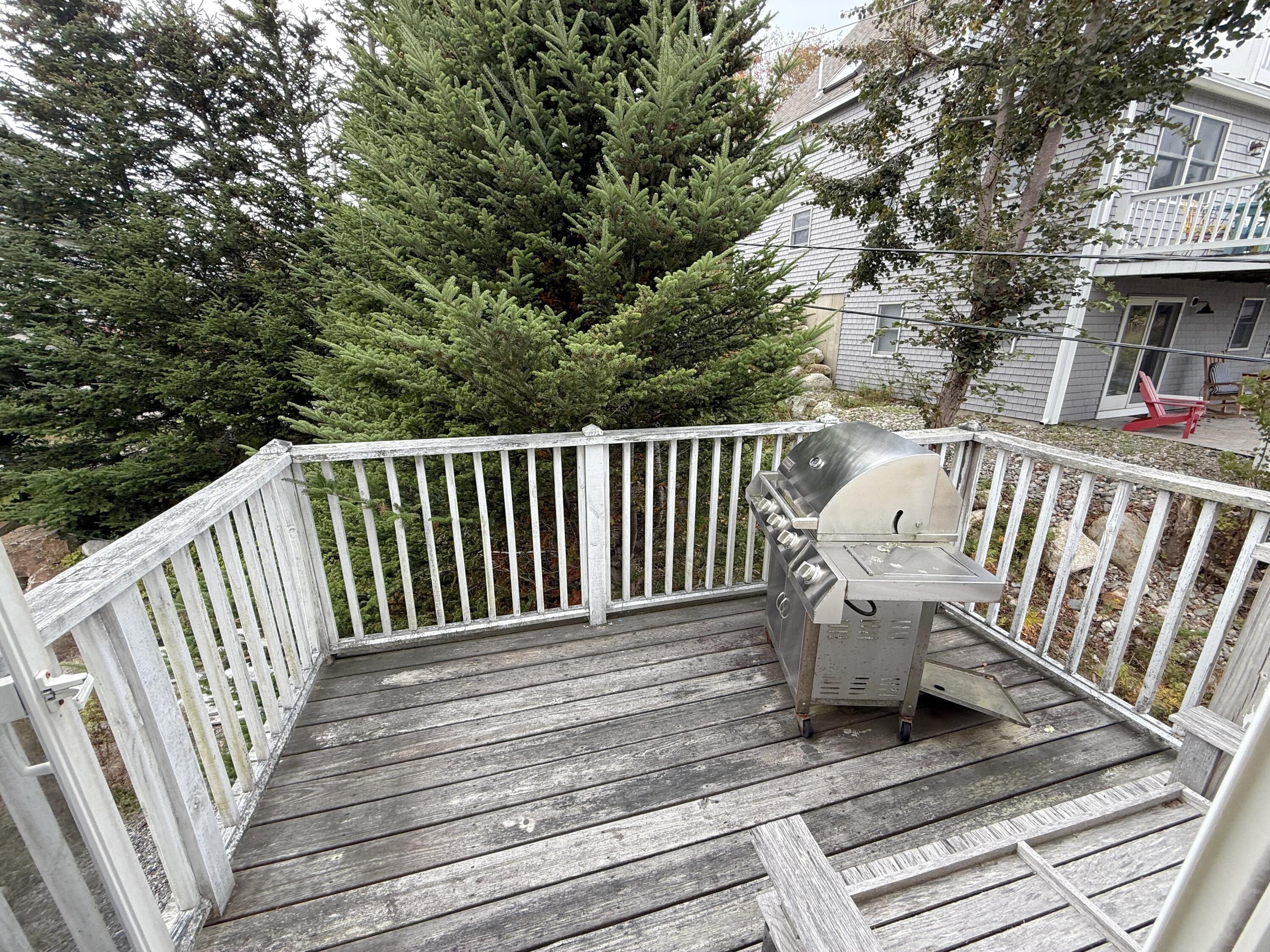 Weathered wooden deck with white railings and a stainless-steel grill, surrounded by evergreen trees.