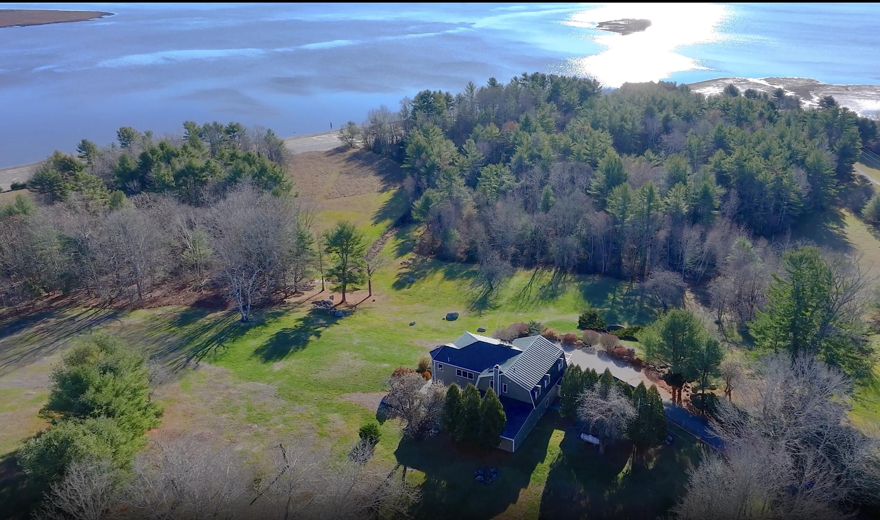 Aerial view of a home on a wooded, open lawn beside a broad body of water.