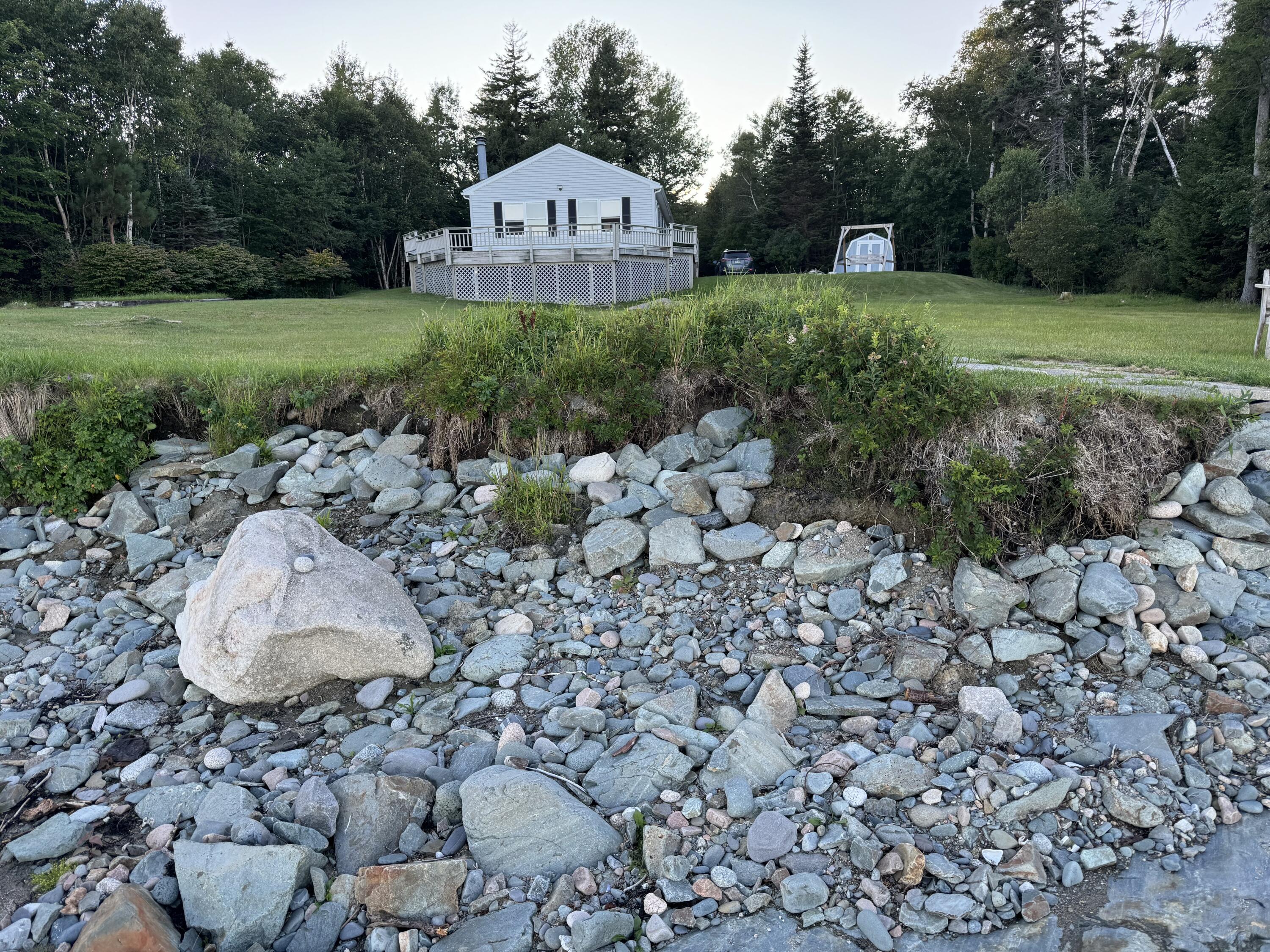 White house with raised deck above a rocky shoreline, surrounded by trees.