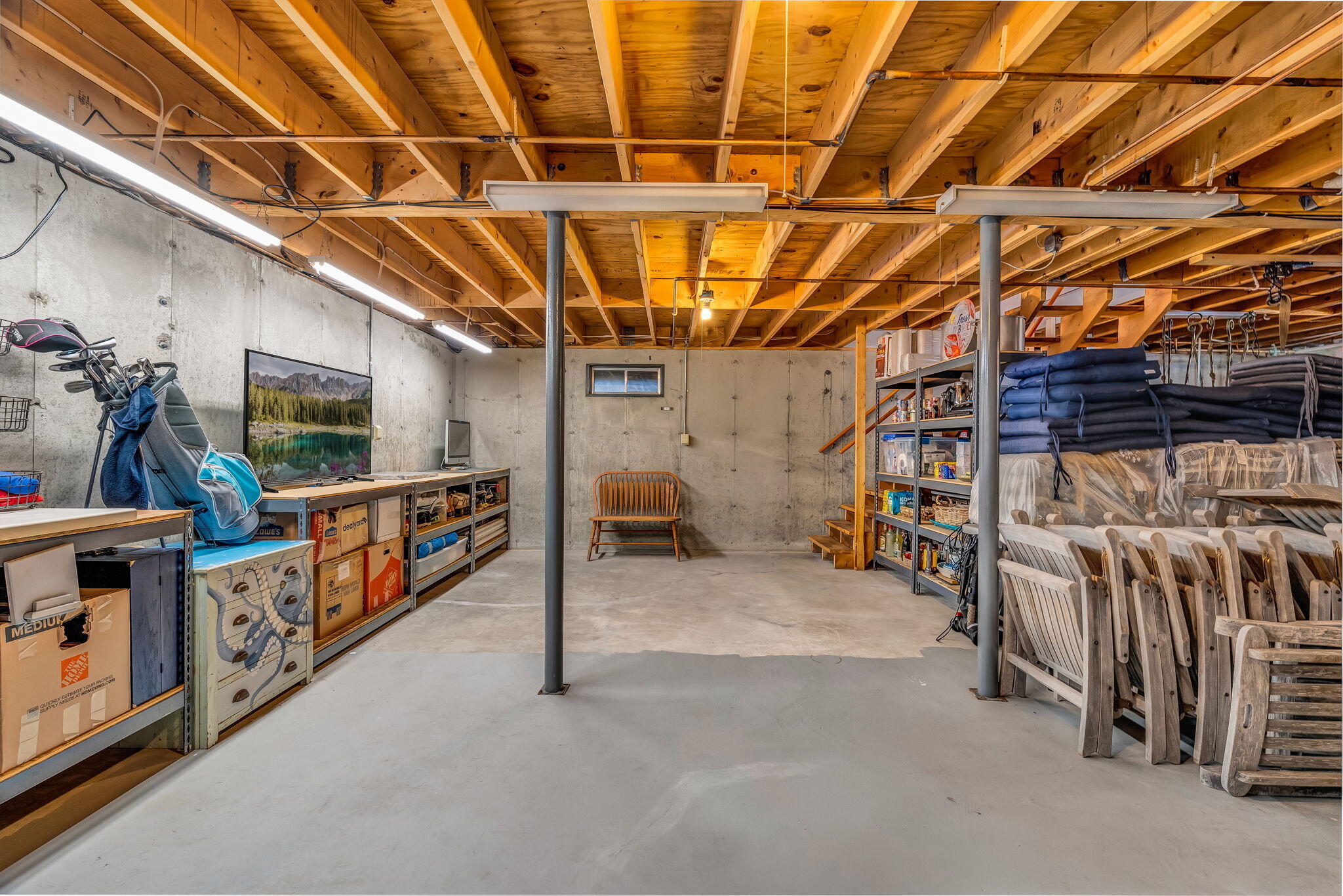 Basement storage area with exposed wood joists, shelving, golf clubs, and stacked patio furniture.