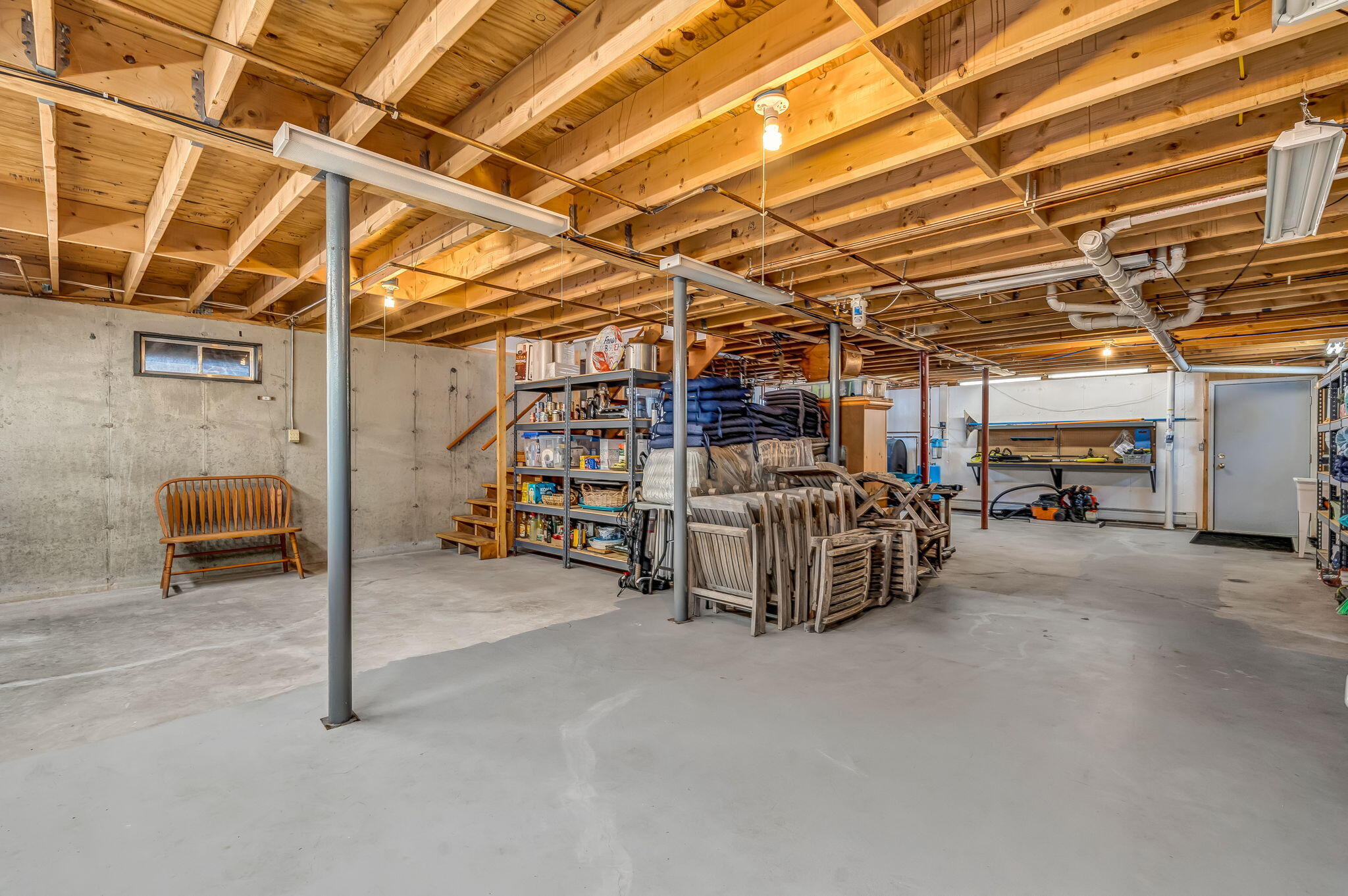 Spacious basement with exposed wooden joists, concrete floors, storage shelves, and stacked patio furniture.