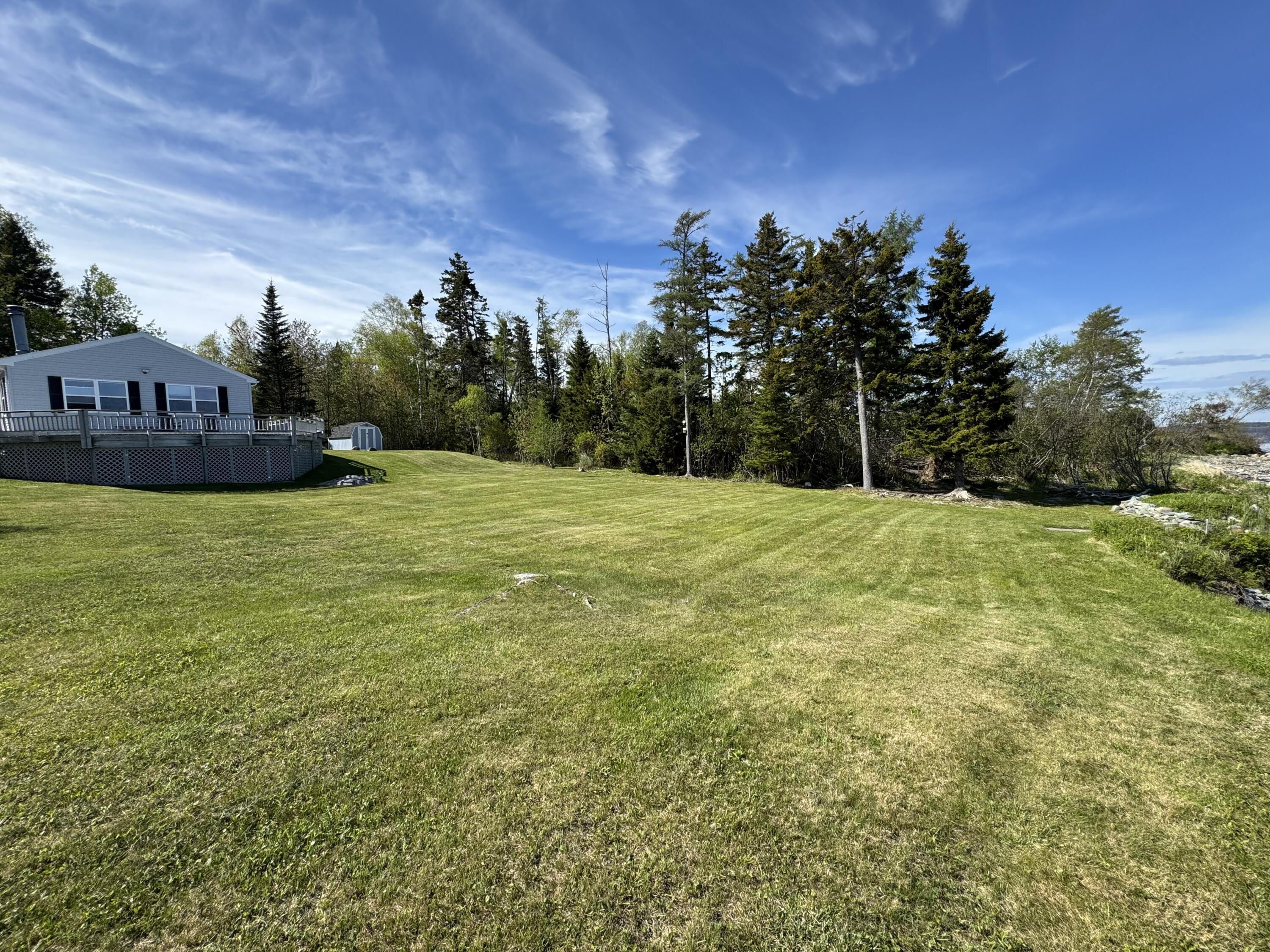 Gray house with raised deck beside a broad lawn, trees, and a glimpse of shoreline water.