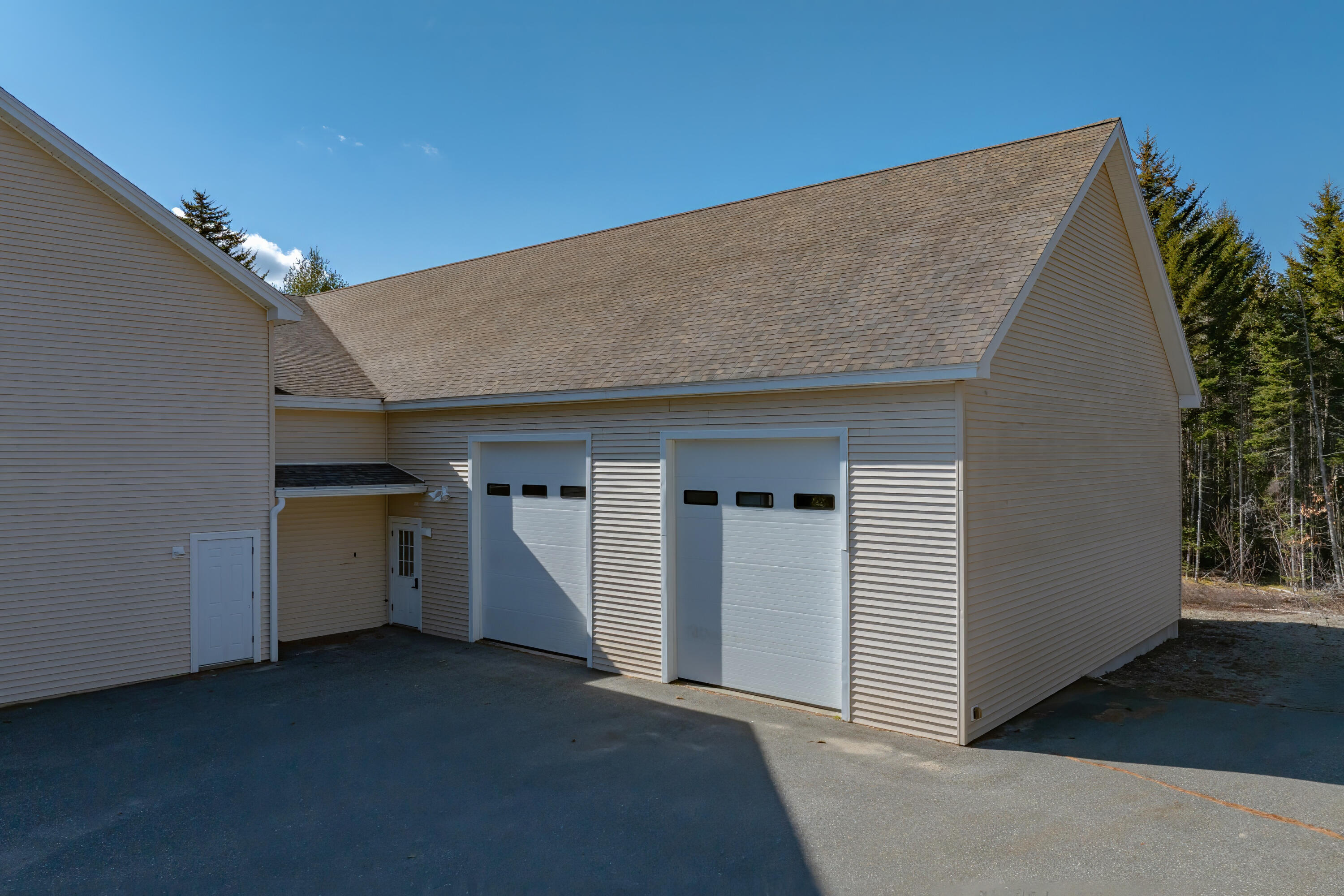 Beige garage building with two white overhead doors beside a paved parking area and evergreen woods.