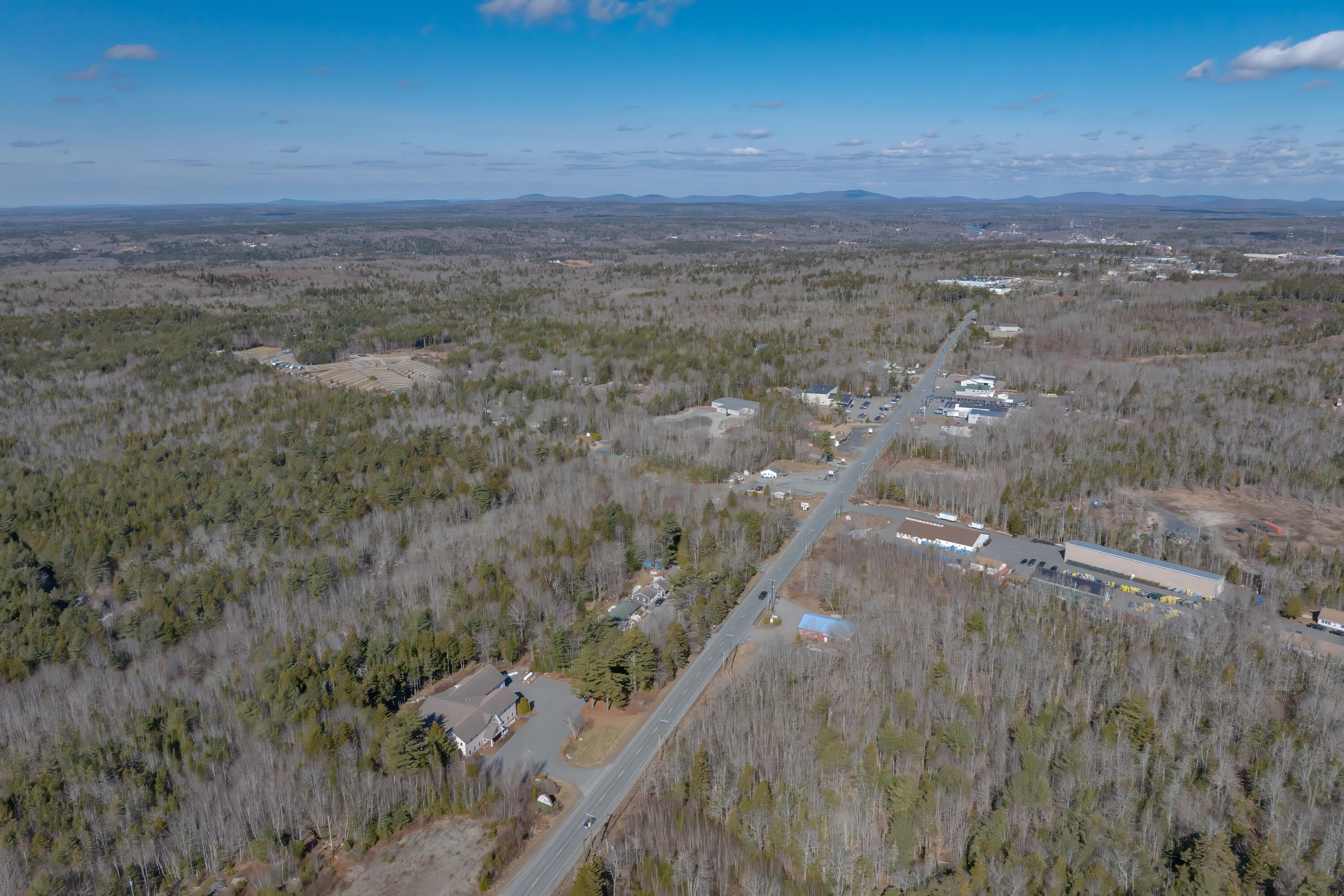 Aerial view of a road winding through wooded land, with scattered buildings and distant hills.