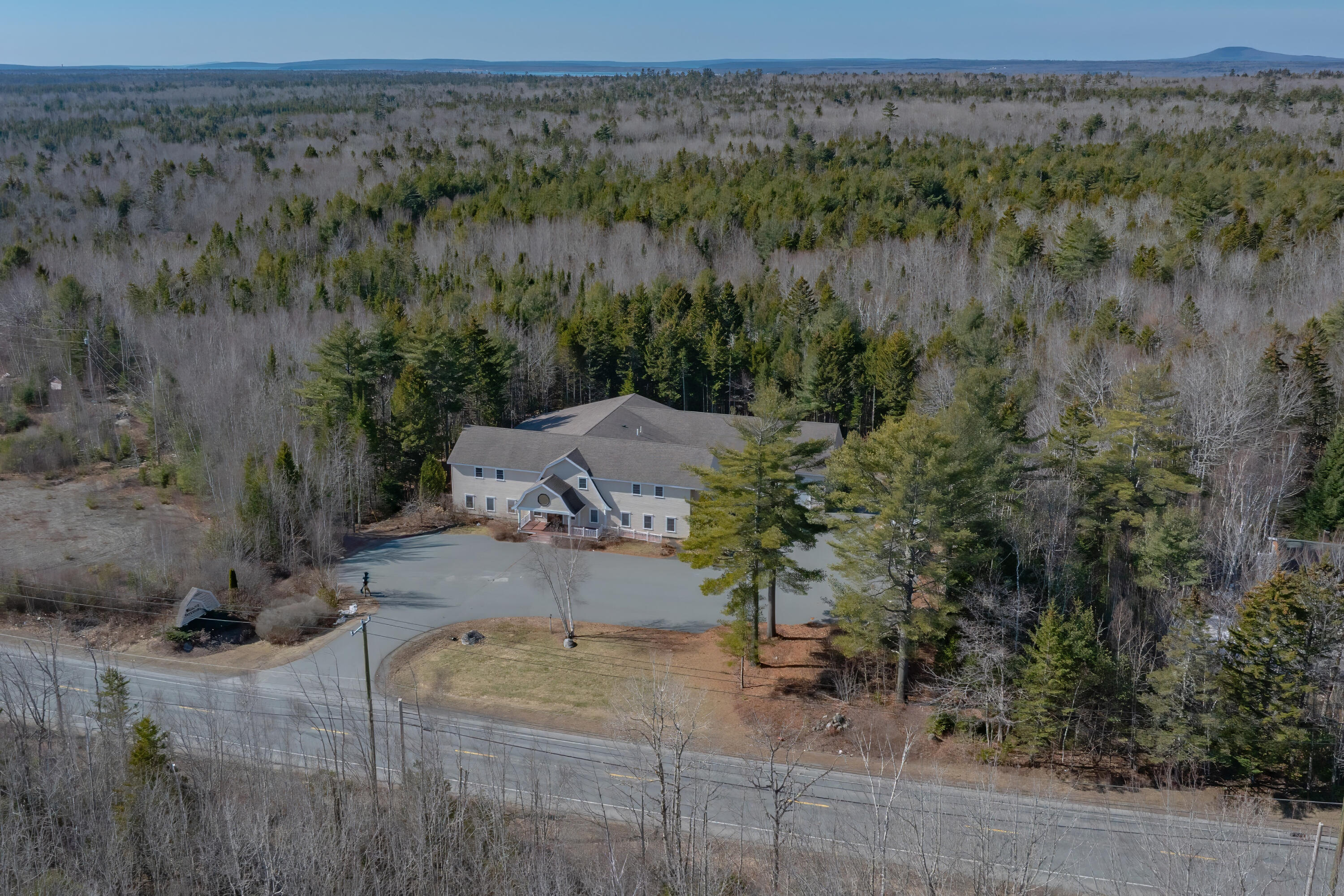 Aerial view of a large building and paved parking area surrounded by woodland beside a road.